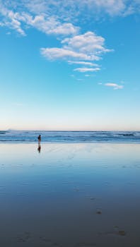 A lone person walks along a serene beach under a clear blue sky at sunrise.