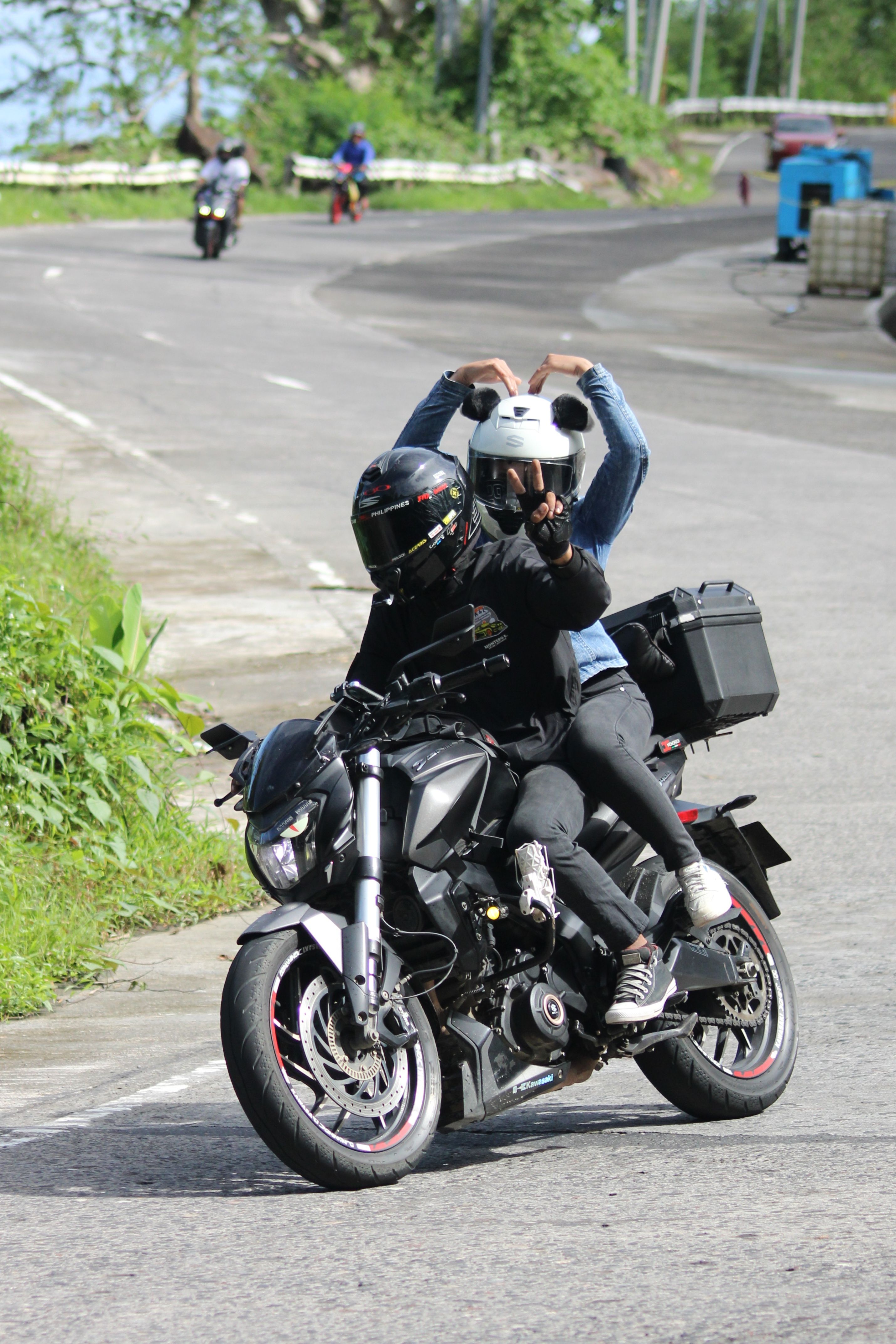 Two motorcyclists enjoy a scenic ride on a winding road, making heart signs.