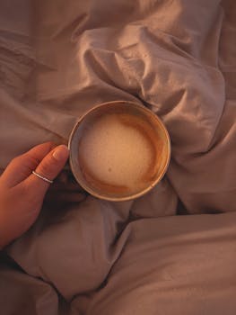 A comforting scene of a hand holding a creamy coffee cup on soft bedding.