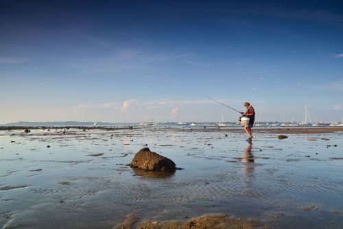 Free A fisherman casts his line on a serene beach during low tide, surrounded by calm seas and distant boats. Stock Photo