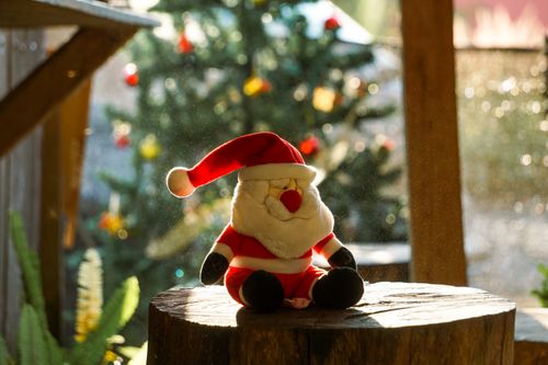 Free A Santa Claus toy sits on a tree stump in the sunlight, with a decorated Christmas tree in the background. Stock Photo