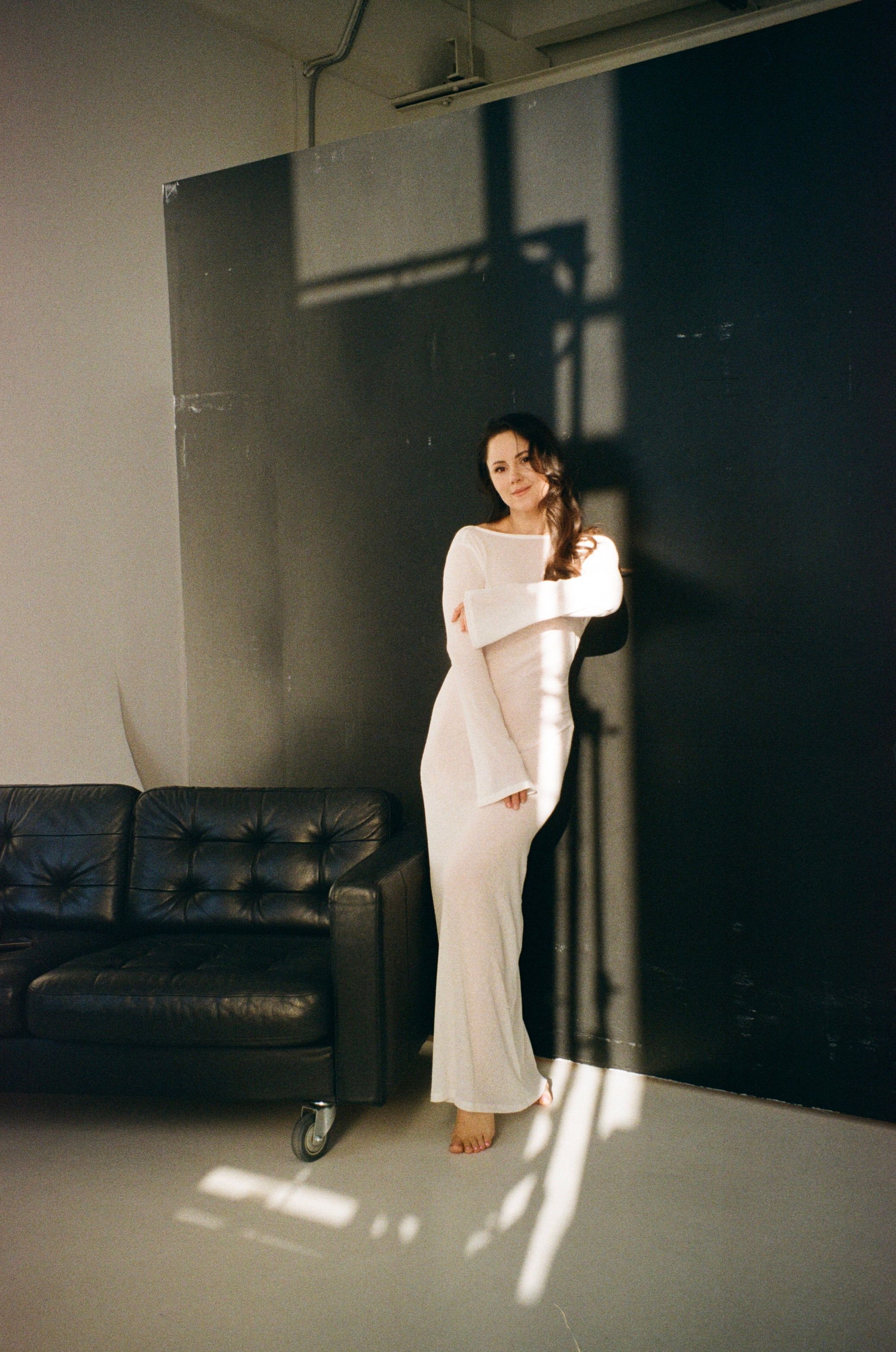 A woman in a white dress stands against a black wall with sunlit shadows indoors.