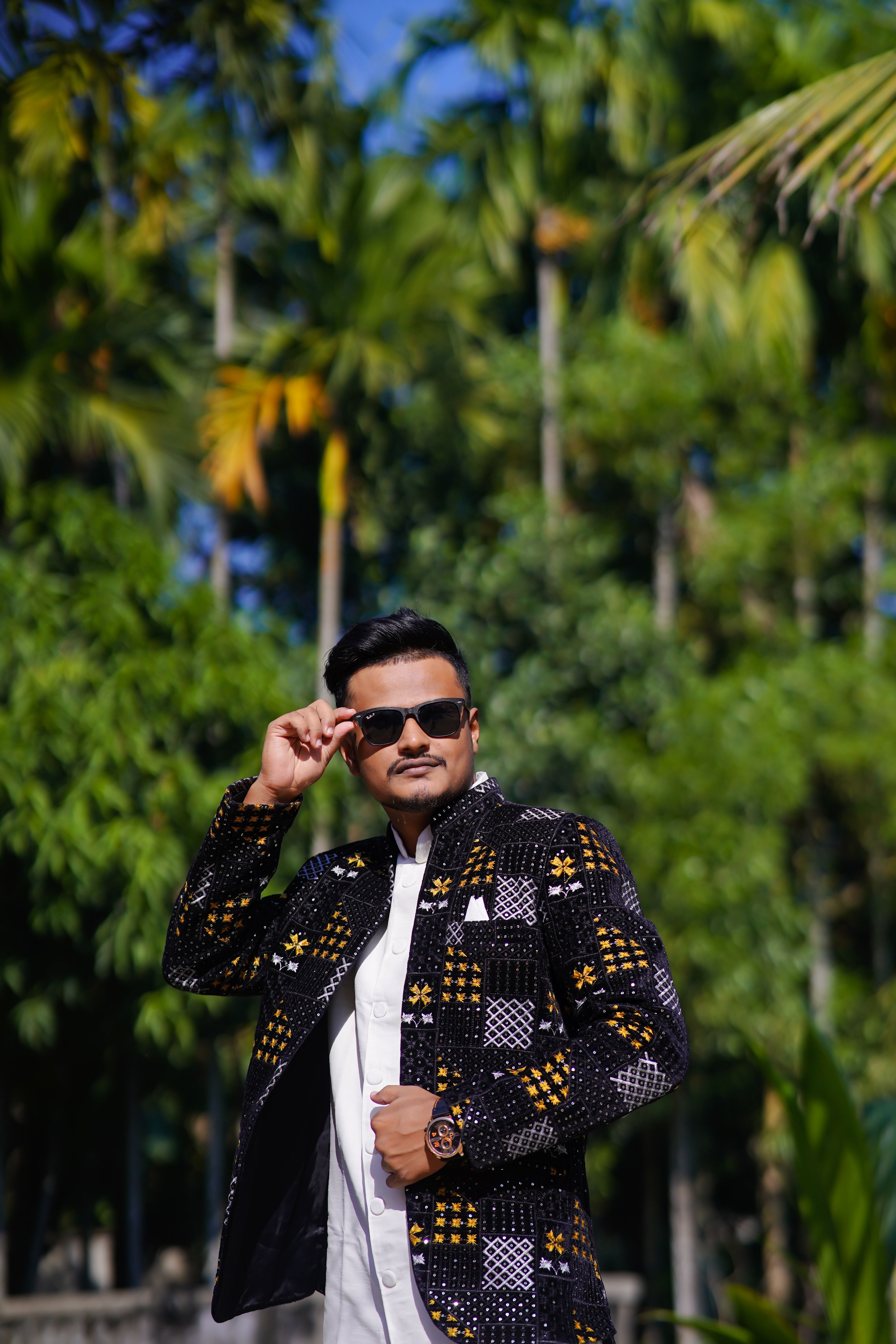 A man wearing sunglasses poses confidently with a stylish jacket in a lush outdoor setting.
