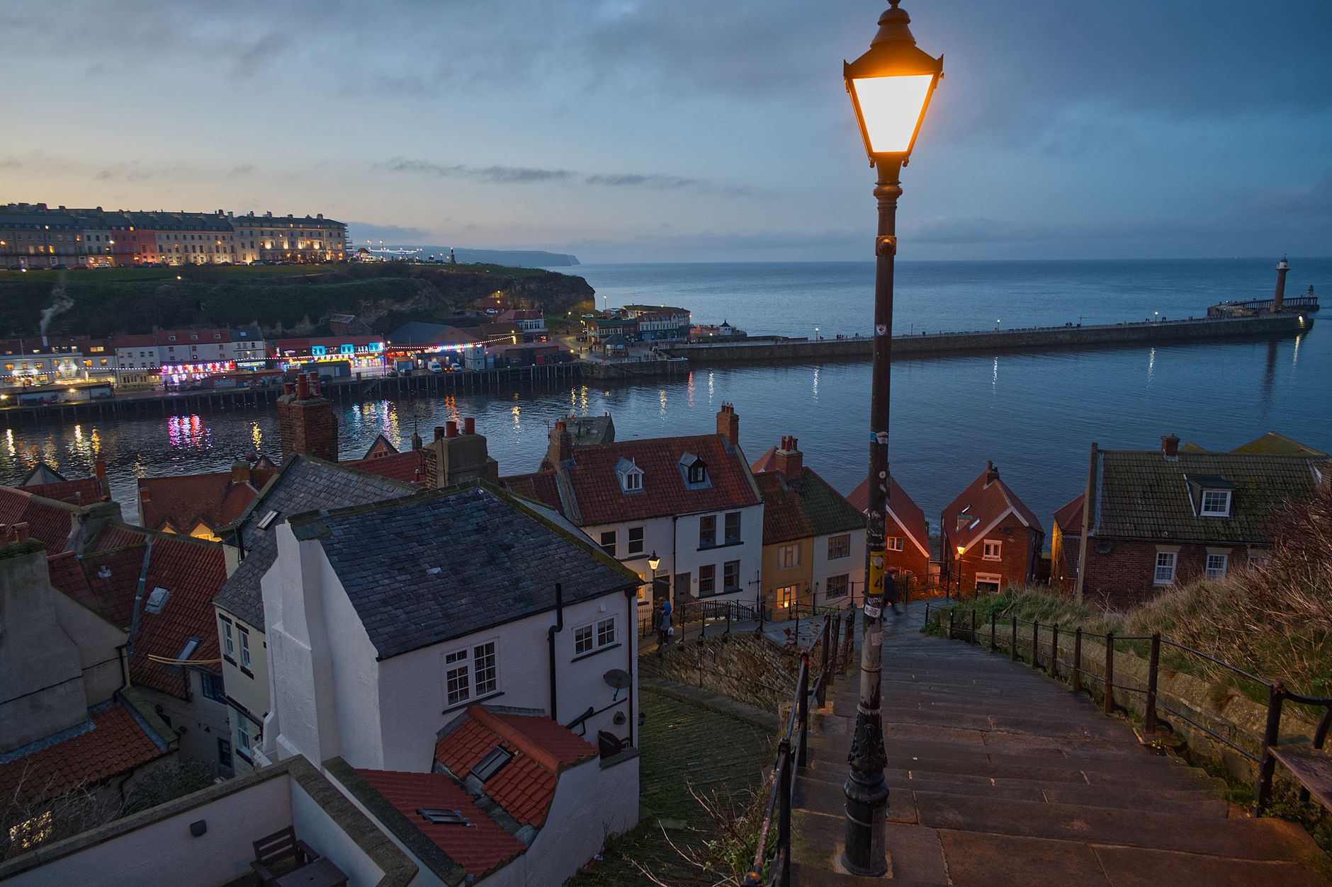 Whitby's 199 steps at dusk, a lit Victorian lamp above the red pantile rooftops of the old town, with the harbour and West Pier beyond.