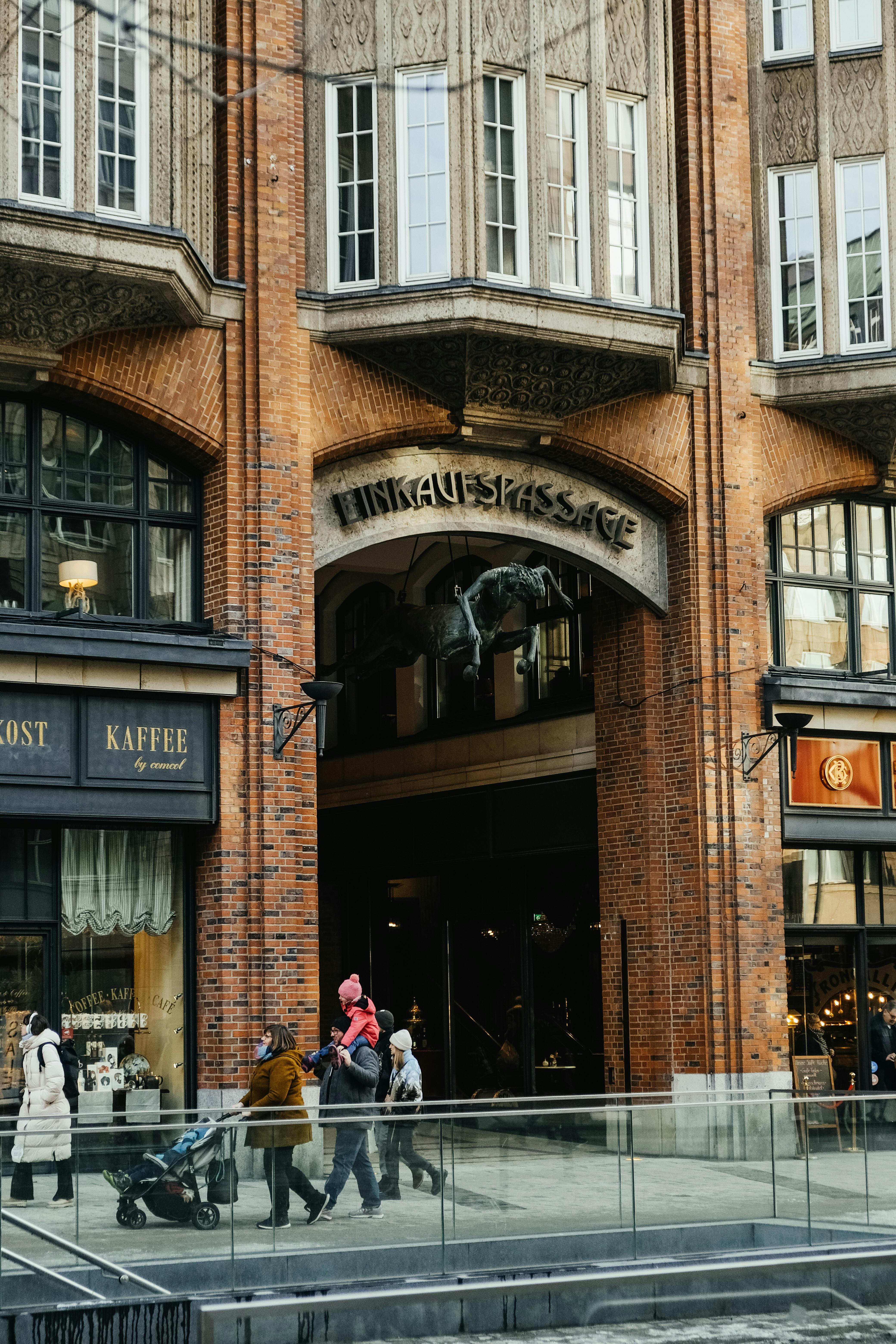 grátis Pessoas caminhando em frente a uma histórica galeria comercial urbana com arquitetura ornamentada em tijolos. Foto profissional