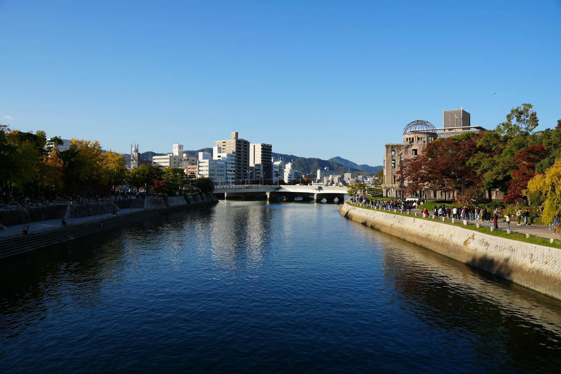 Beautiful autumn scene of Hiroshima Peace Memorial Park with vibrant trees and clear river reflecting the cityscape.