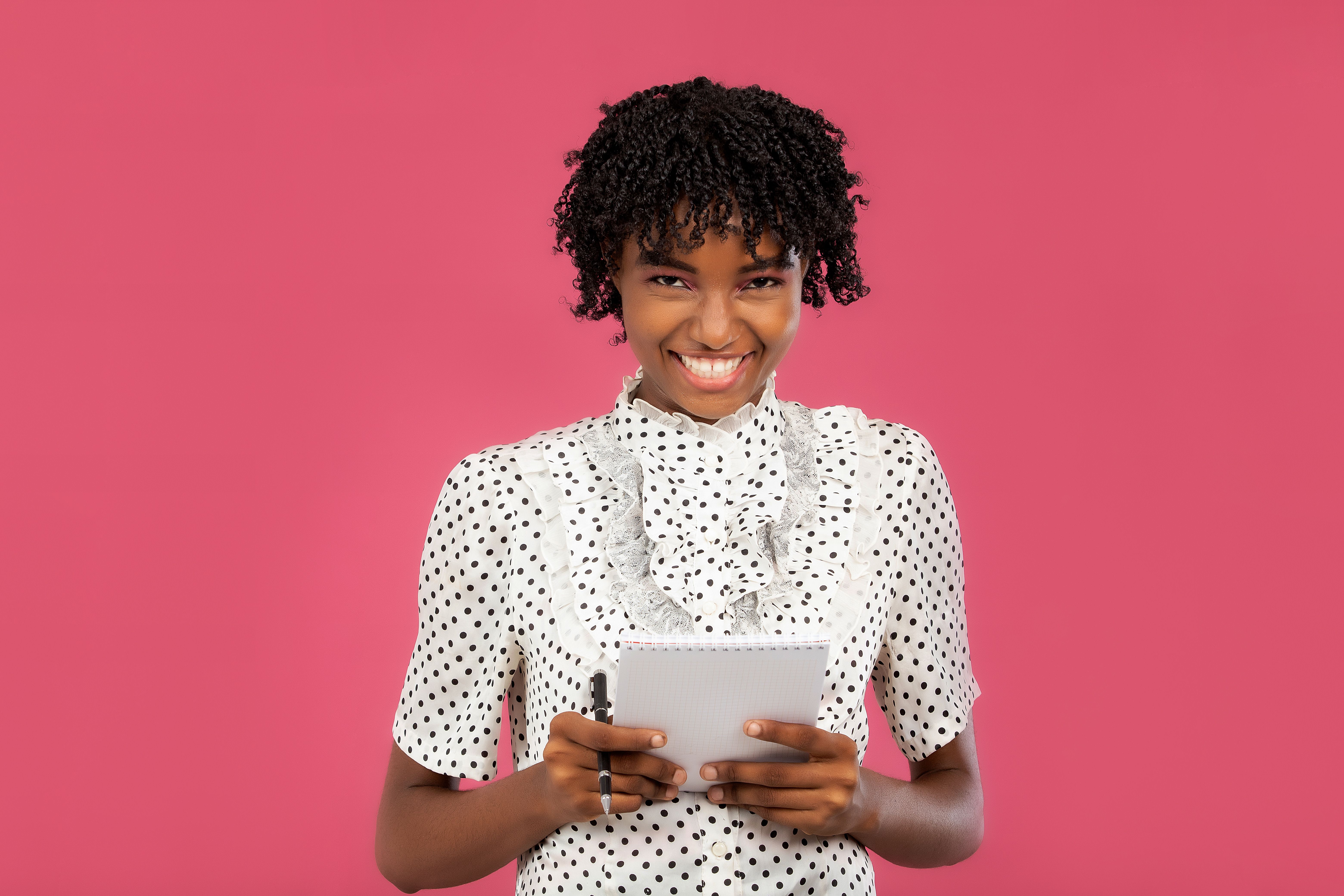 Cheerful woman with curly hair holding a notebook, smiling against a pink background.