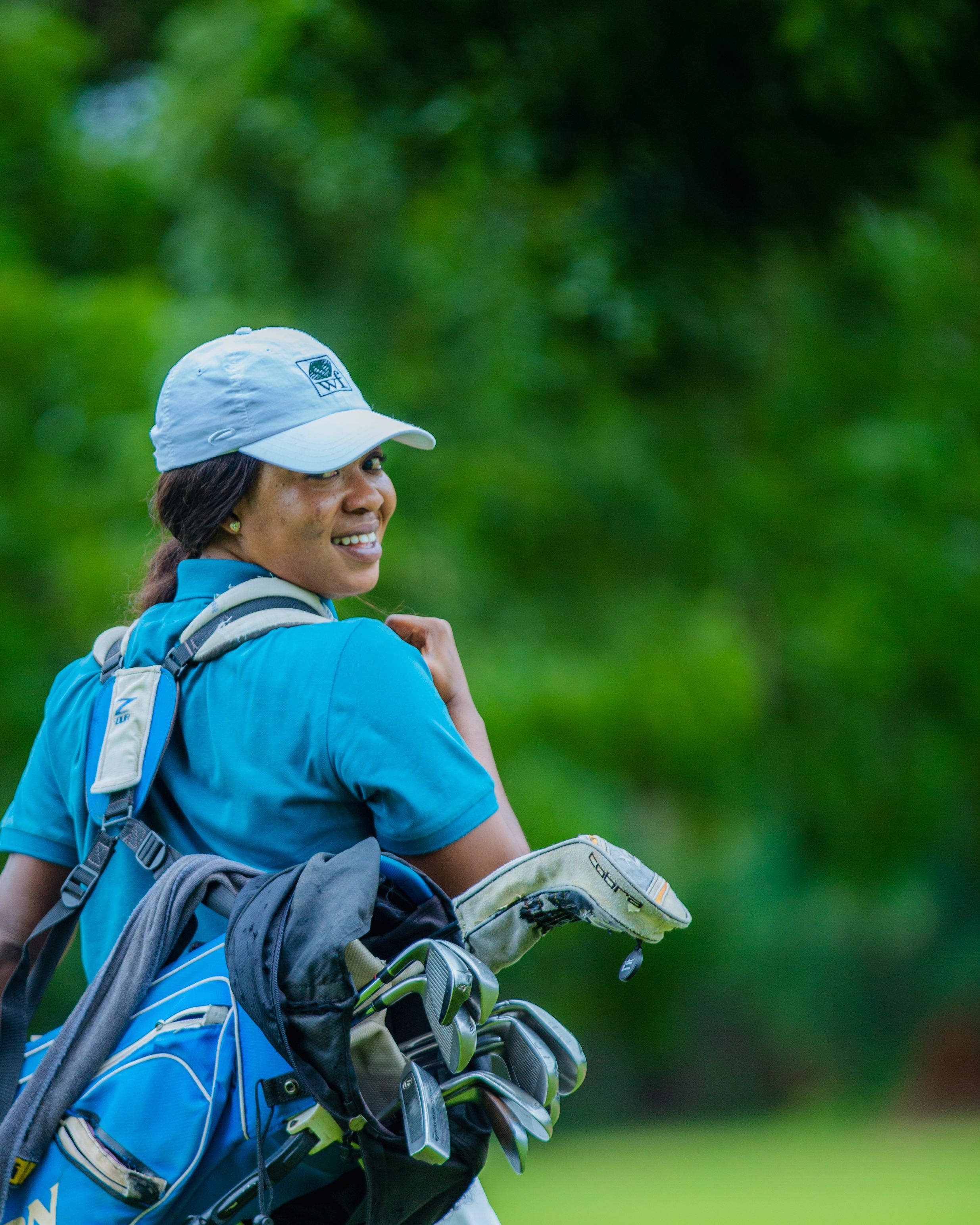 Smiling female golfer with clubs on a lush green course, under bright daylight.