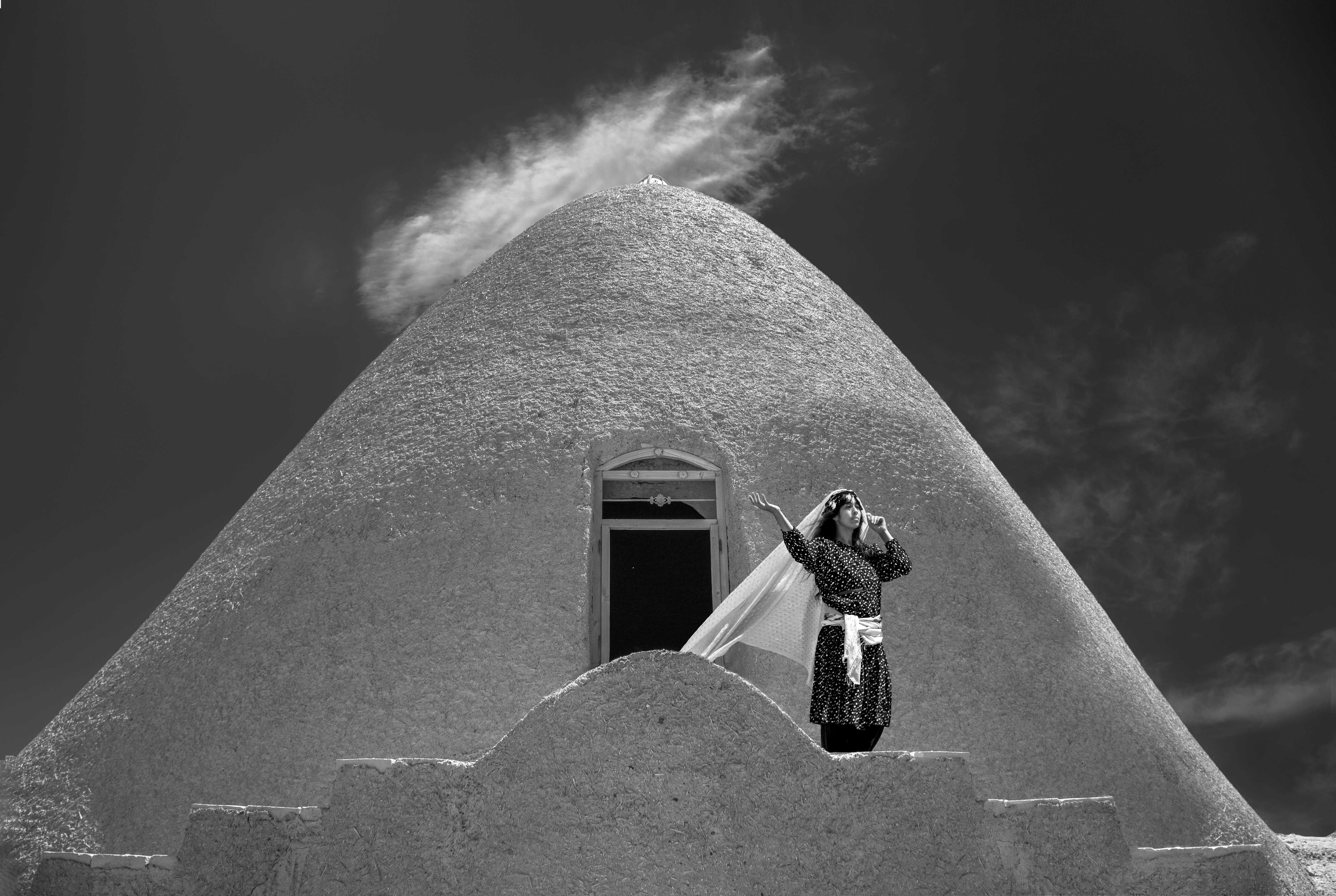 Striking black and white image of a woman posing by a unique cone-shaped structure, under a dynamic sky.