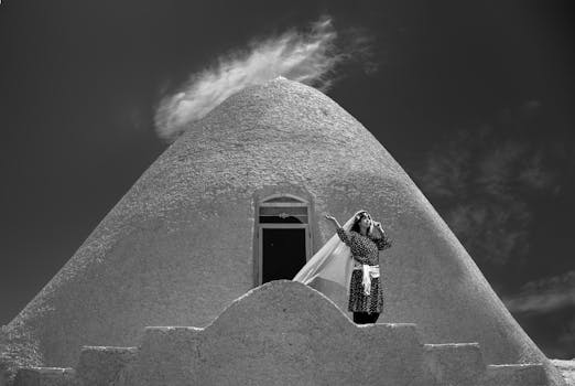 Striking black and white image of a woman posing by a unique cone-shaped structure, under a dynamic sky.