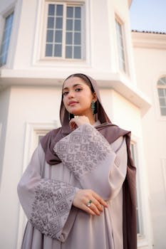 Elegant woman in a patterned hijab and modest dress standing outside a white building at sunset.