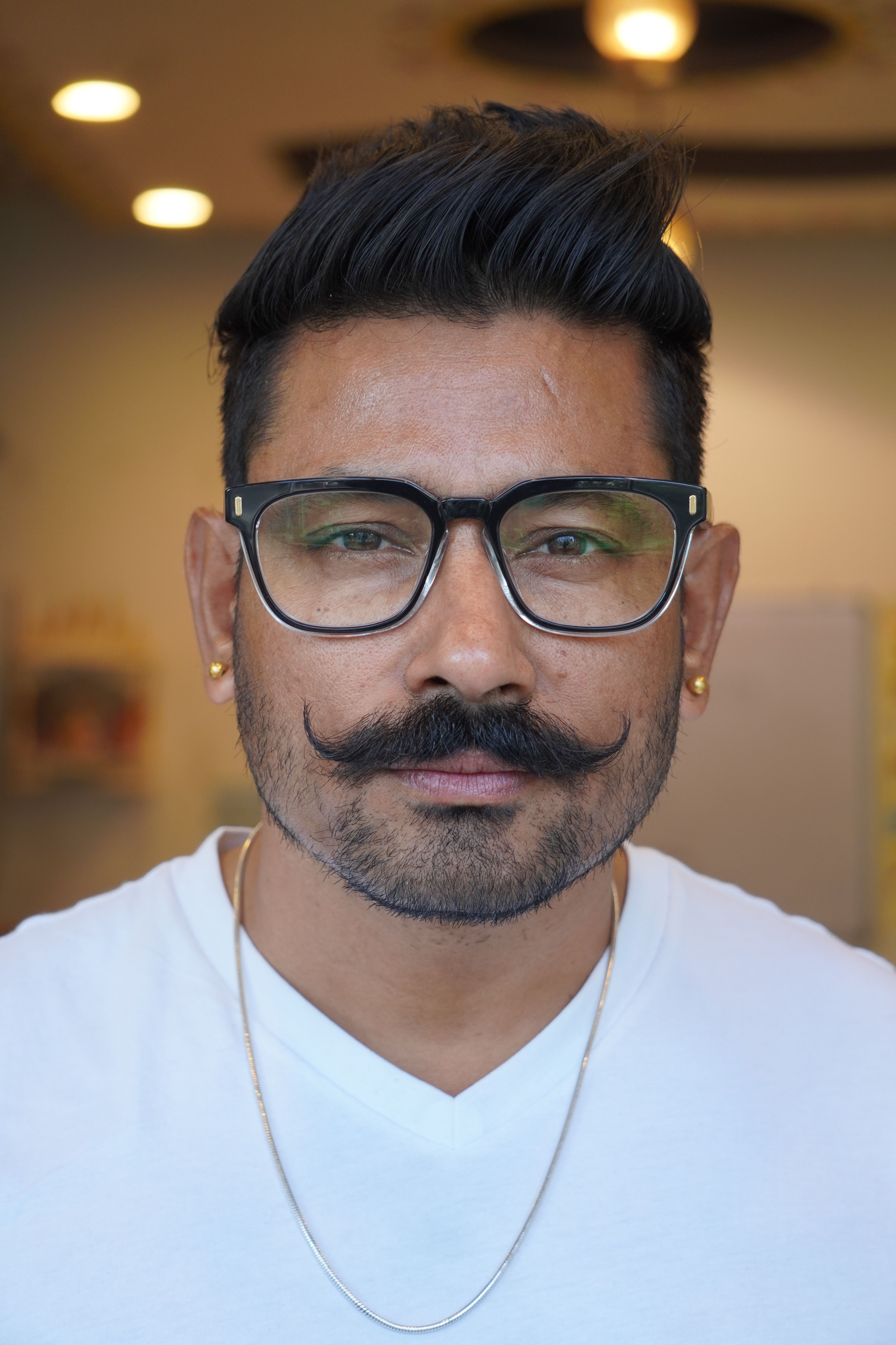 Portrait of a man with stylish glasses, mustache, and necklace indoors.
