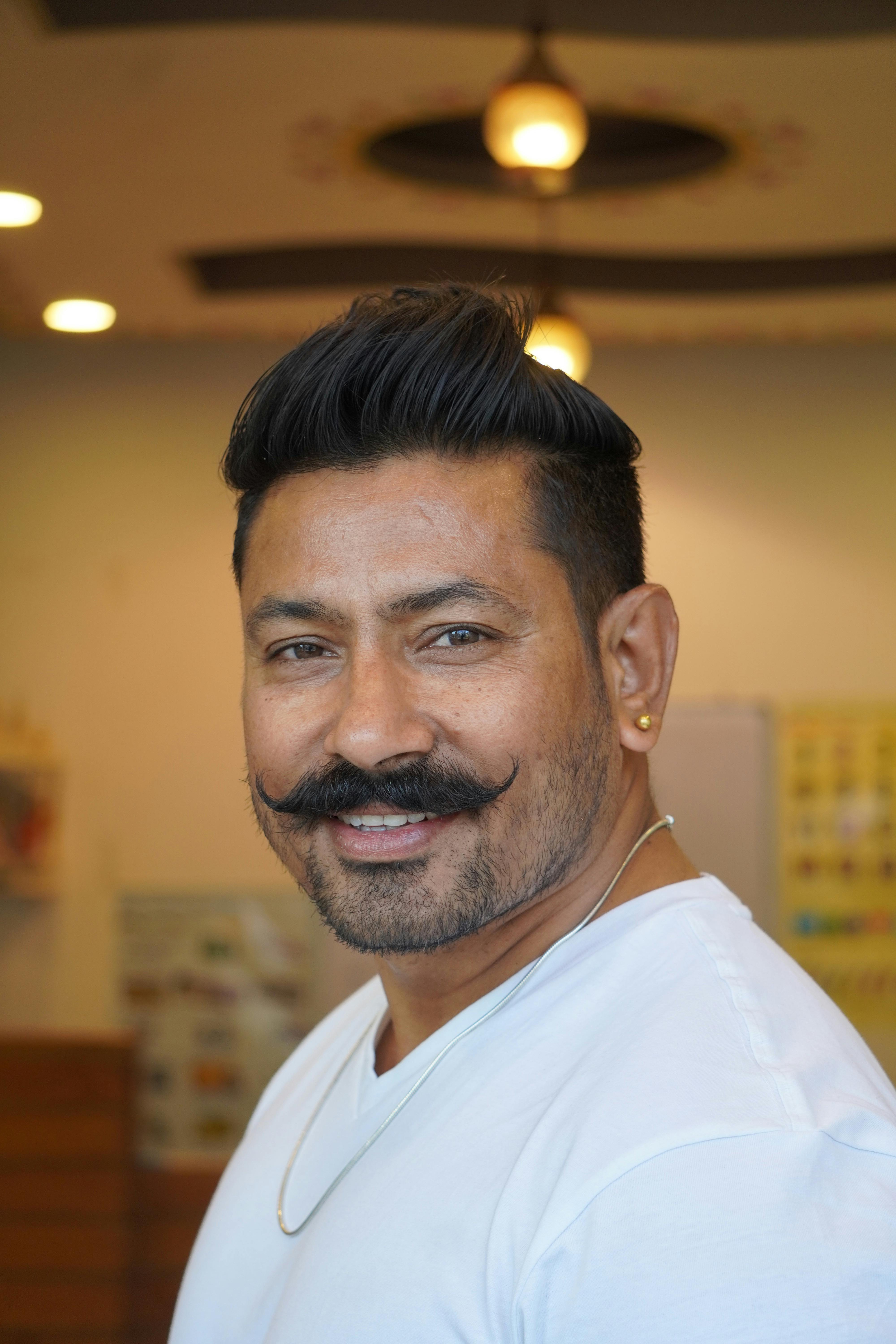 Portrait of a smiling man indoors with a stylish mustache and earrings in a casual white shirt.