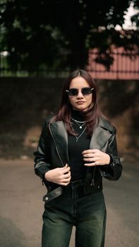 A fashionable woman in a black leather jacket and sunglasses poses confidently outdoors.