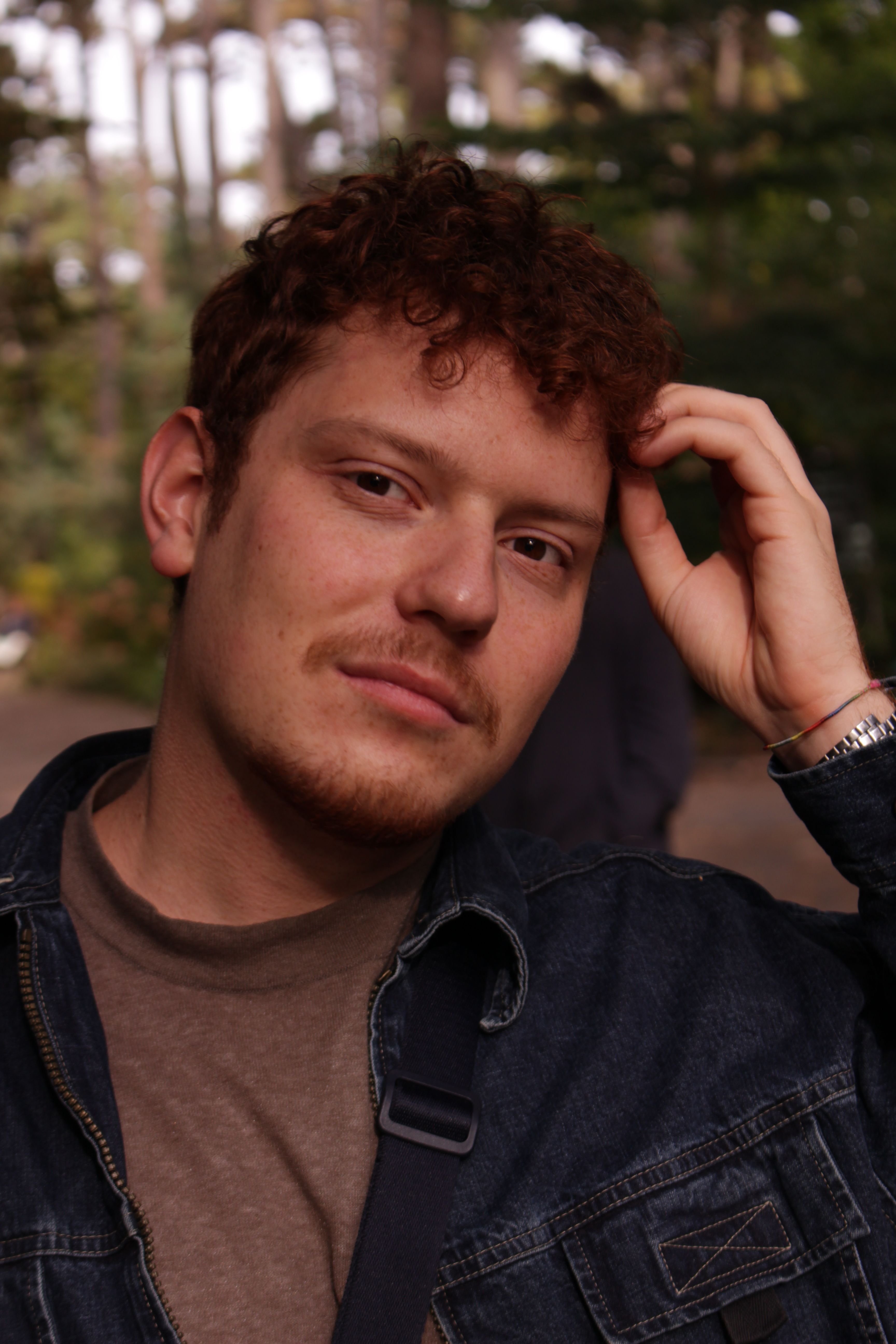 Casual portrait of a man with curly hair wearing a denim jacket, set against an outdoor forest background.