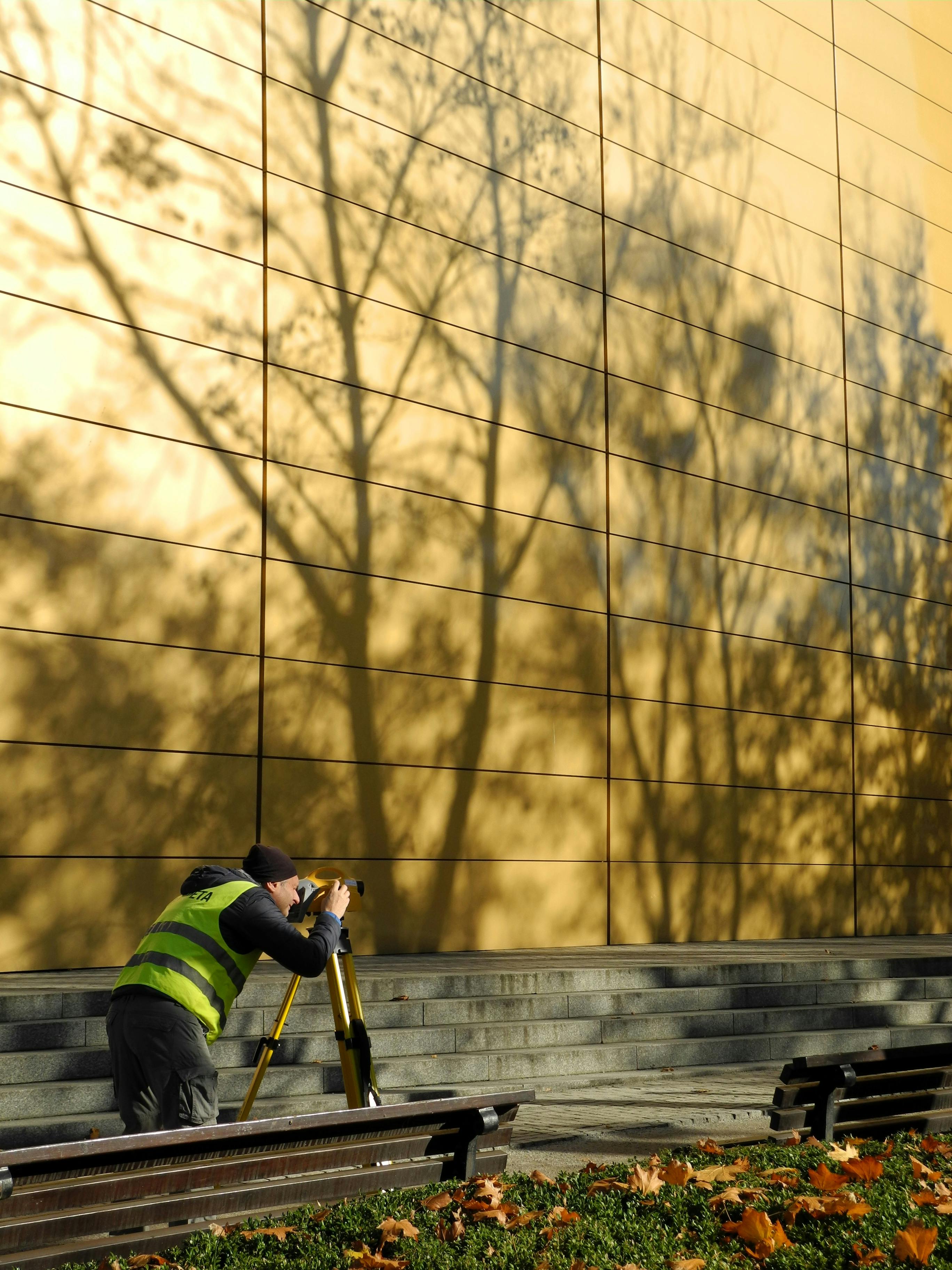 Surveyor in hi-vis vest using equipment near a reflective gold wall with tree shadows.