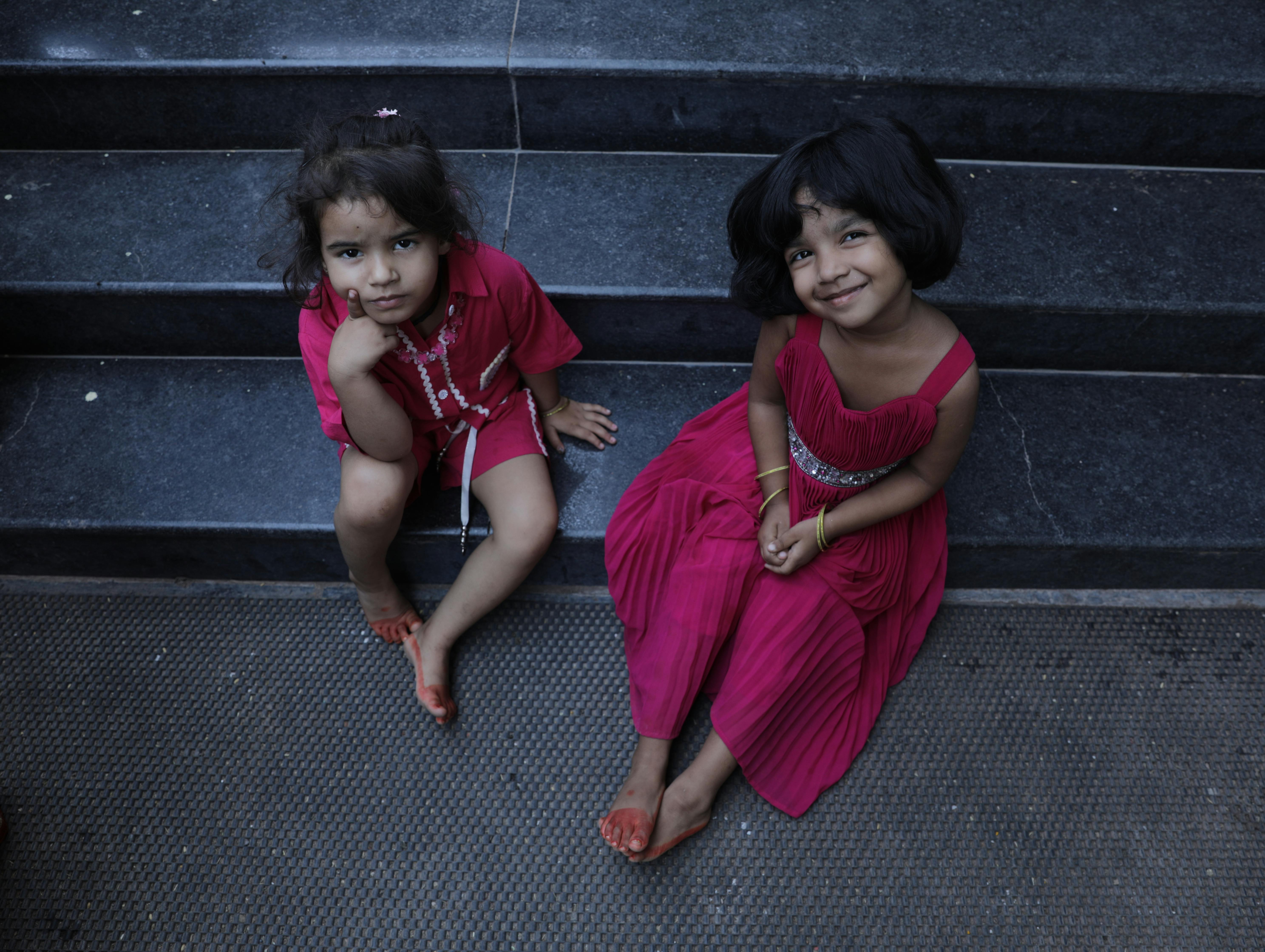 Two charming Indian girls in red dresses smiling while sitting on stairs, capturing childhood joy.