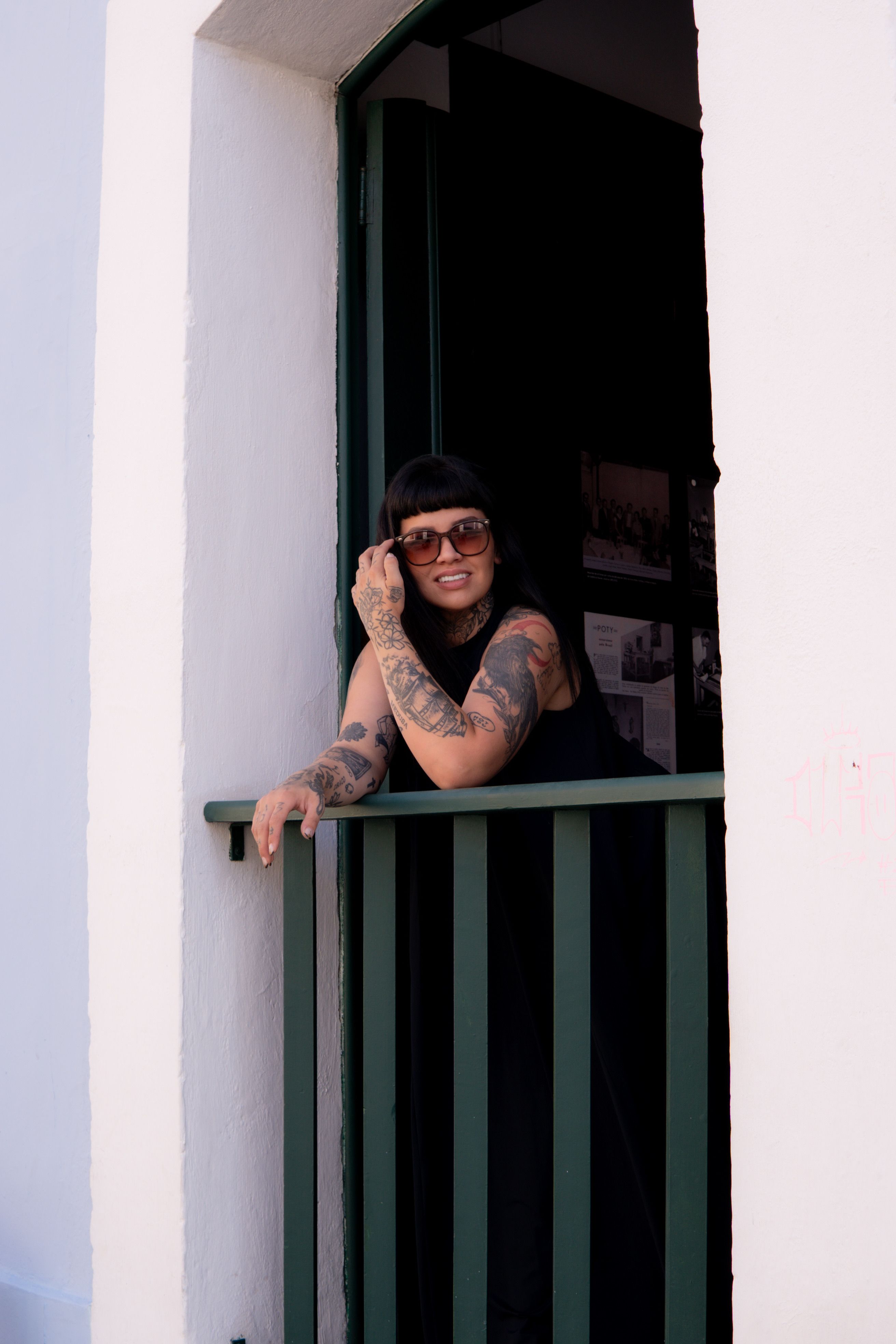 Stylish tattooed woman wearing sunglasses leans on a balcony railing in a relaxed pose.