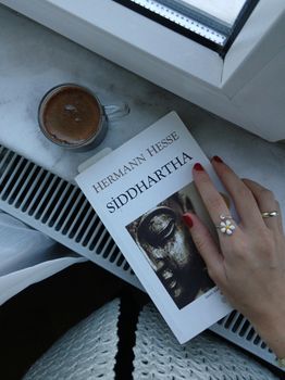 Hand with red nails holding Siddhartha book by window with coffee.