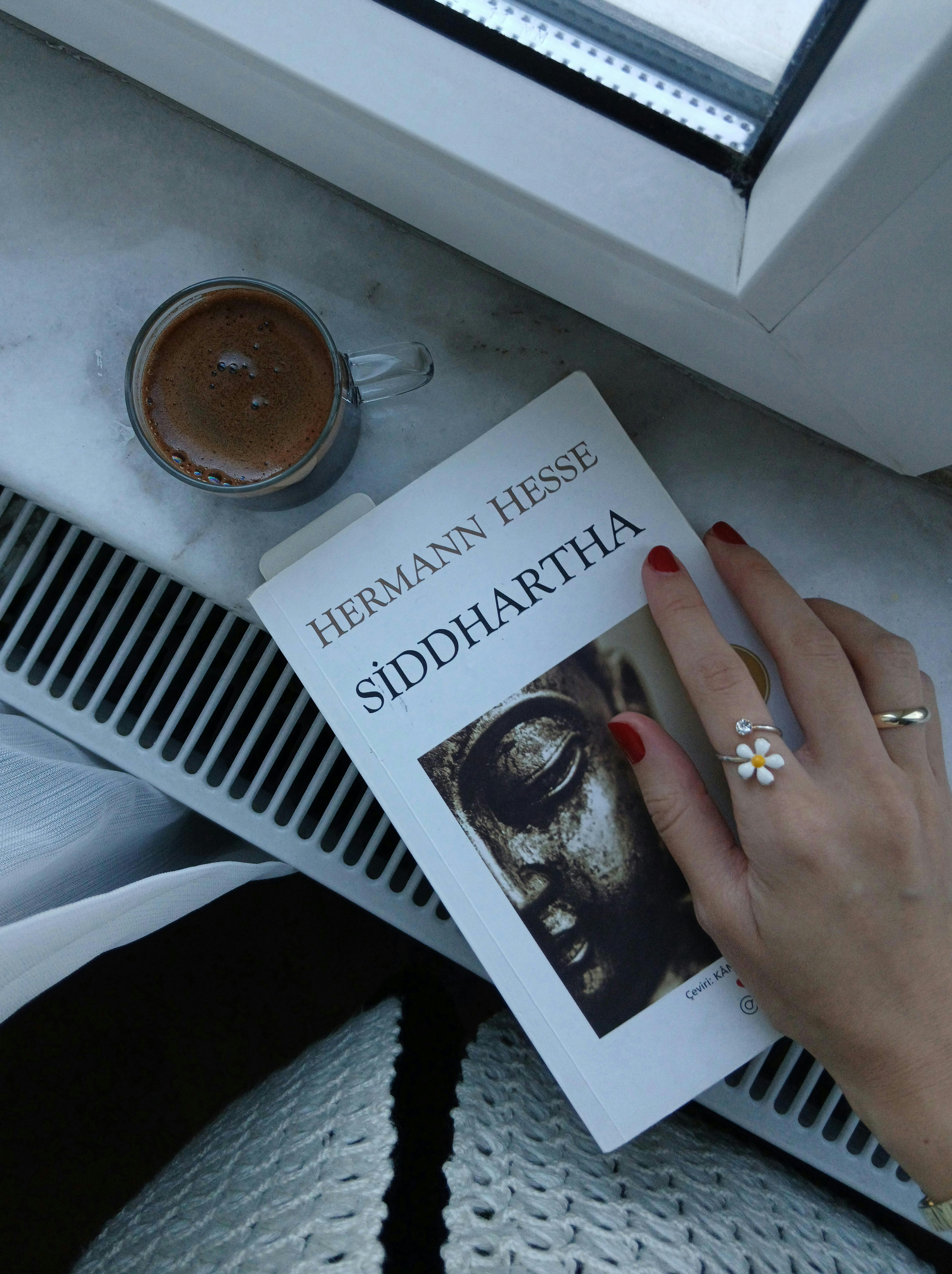 De franc Mà amb ungles vermelles sostenint el llibre de Siddhartha per la finestra amb cafè. Foto d'estoc