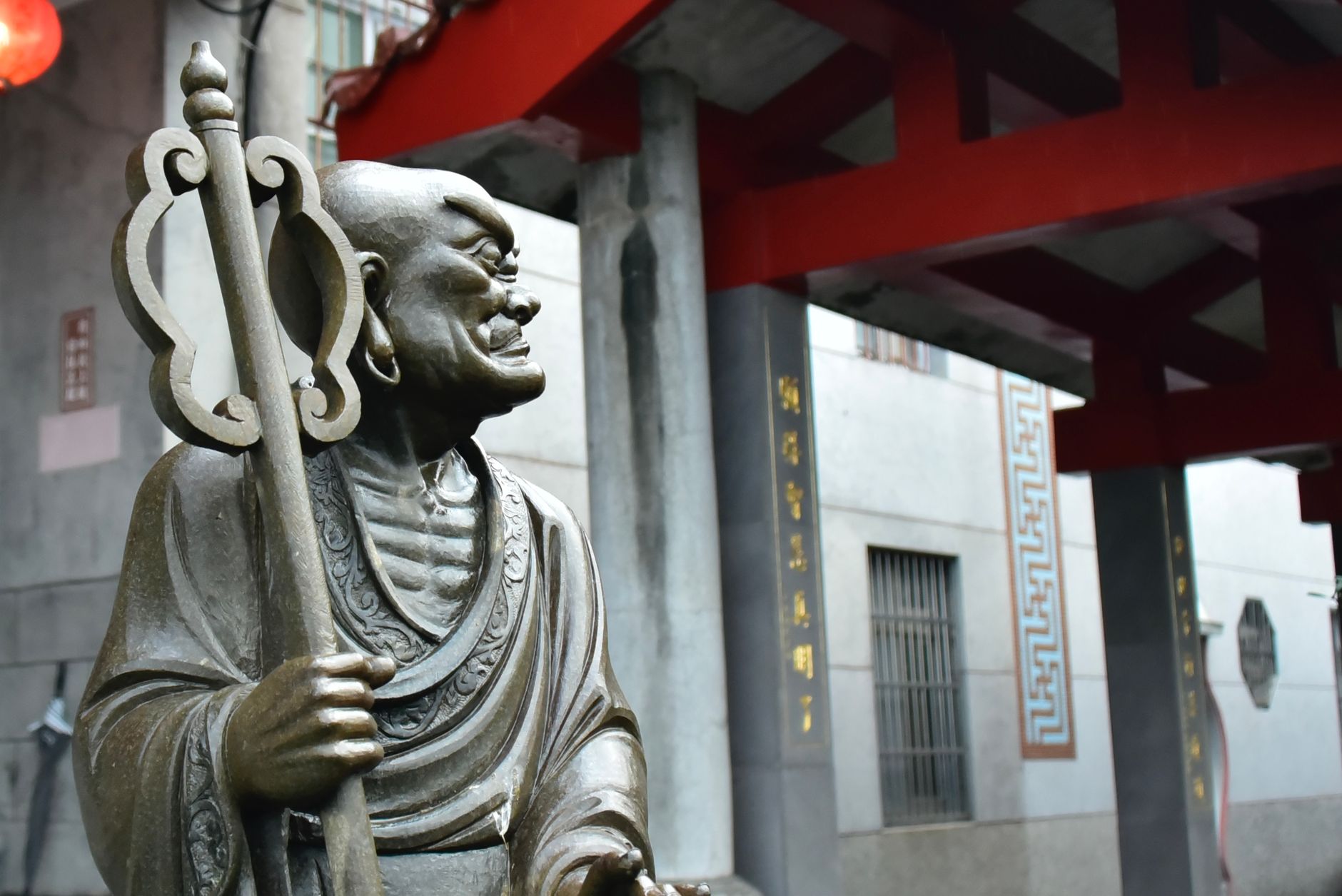 Bronze statue depicting a Buddhist monk with staff, set in a traditional temple courtyard.
