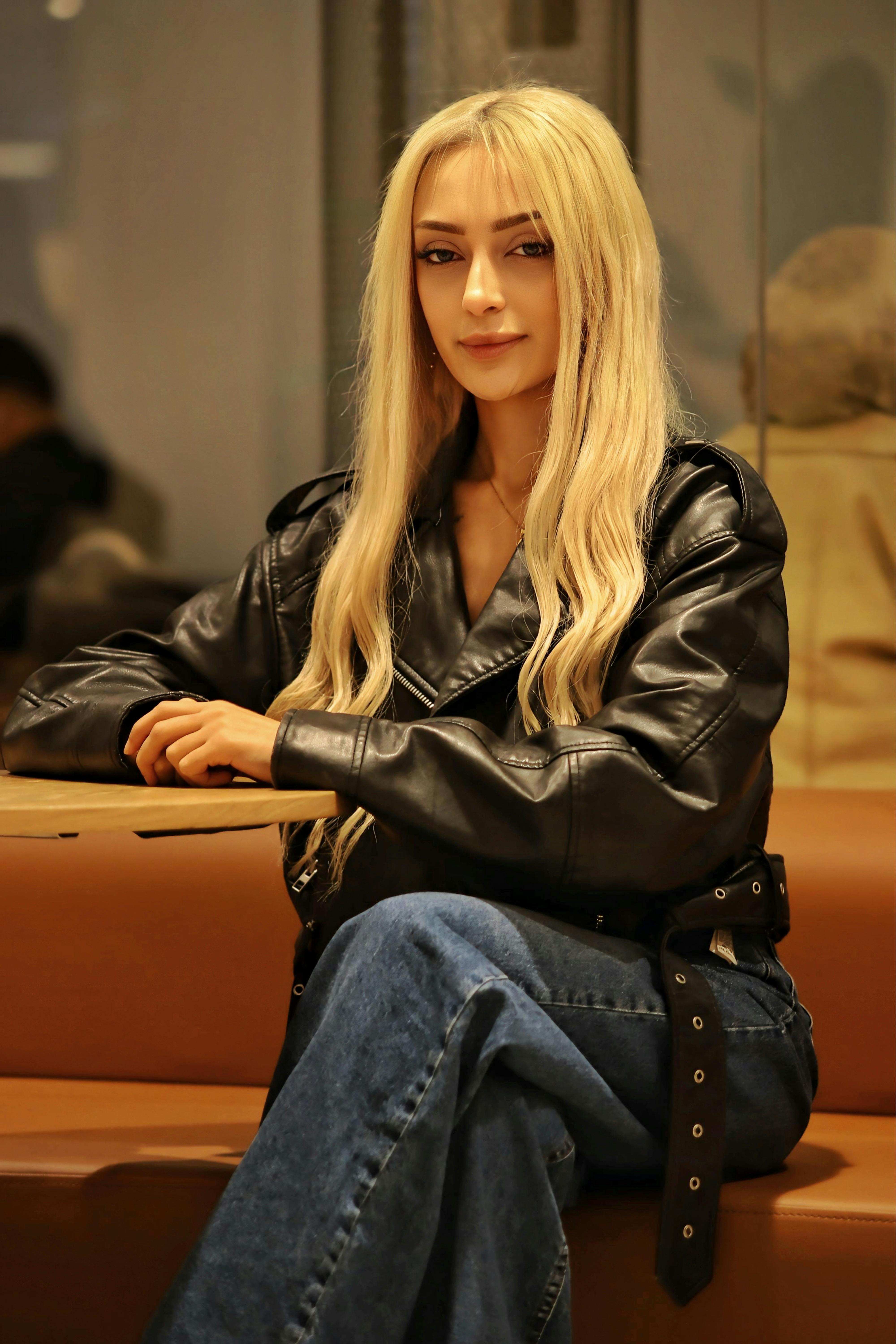 A fashionable woman sits in a cafe, wearing a black leather jacket and jeans, exuding confidence.