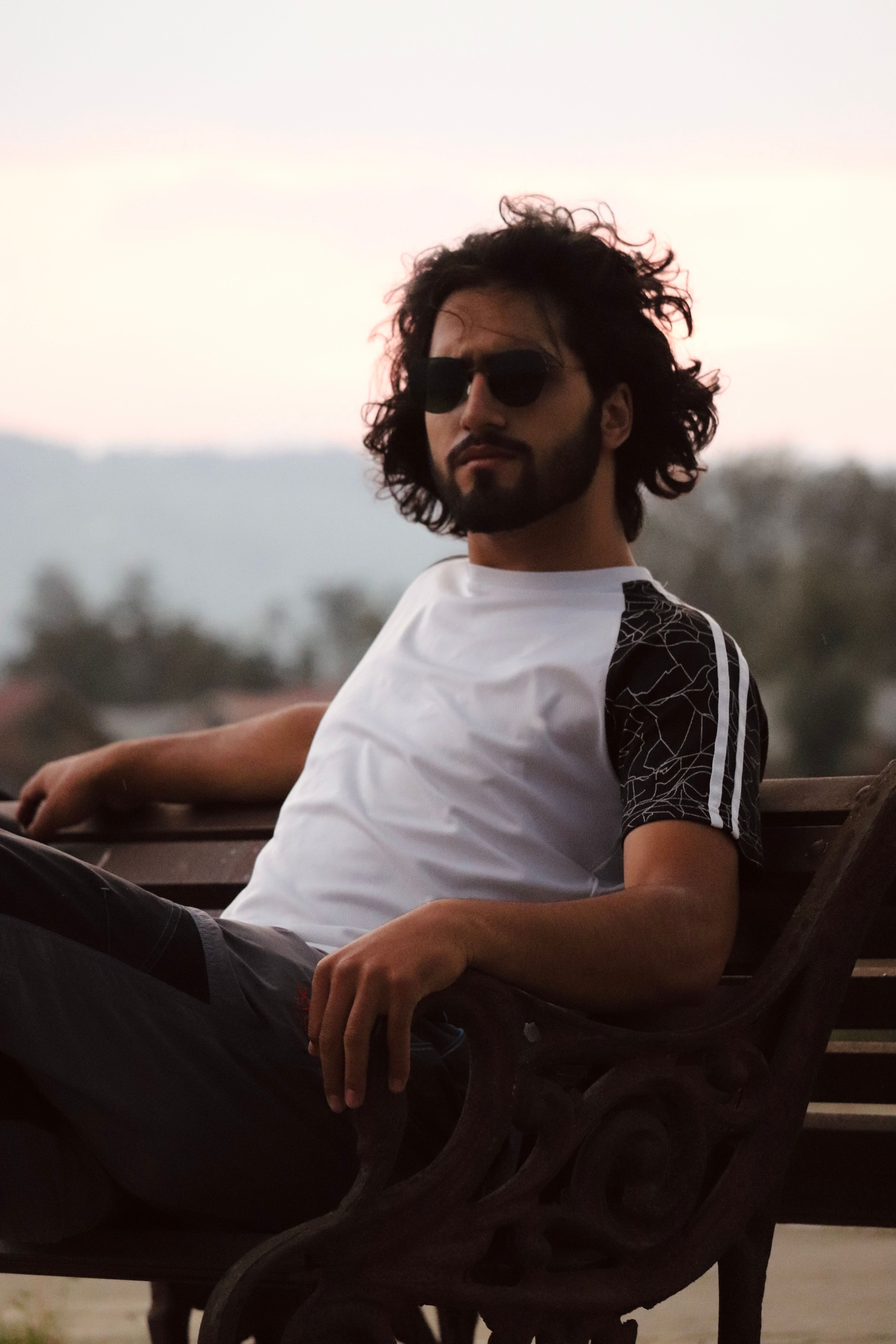 A man in sunglasses and casual attire relaxes on an outdoor bench at sunset.