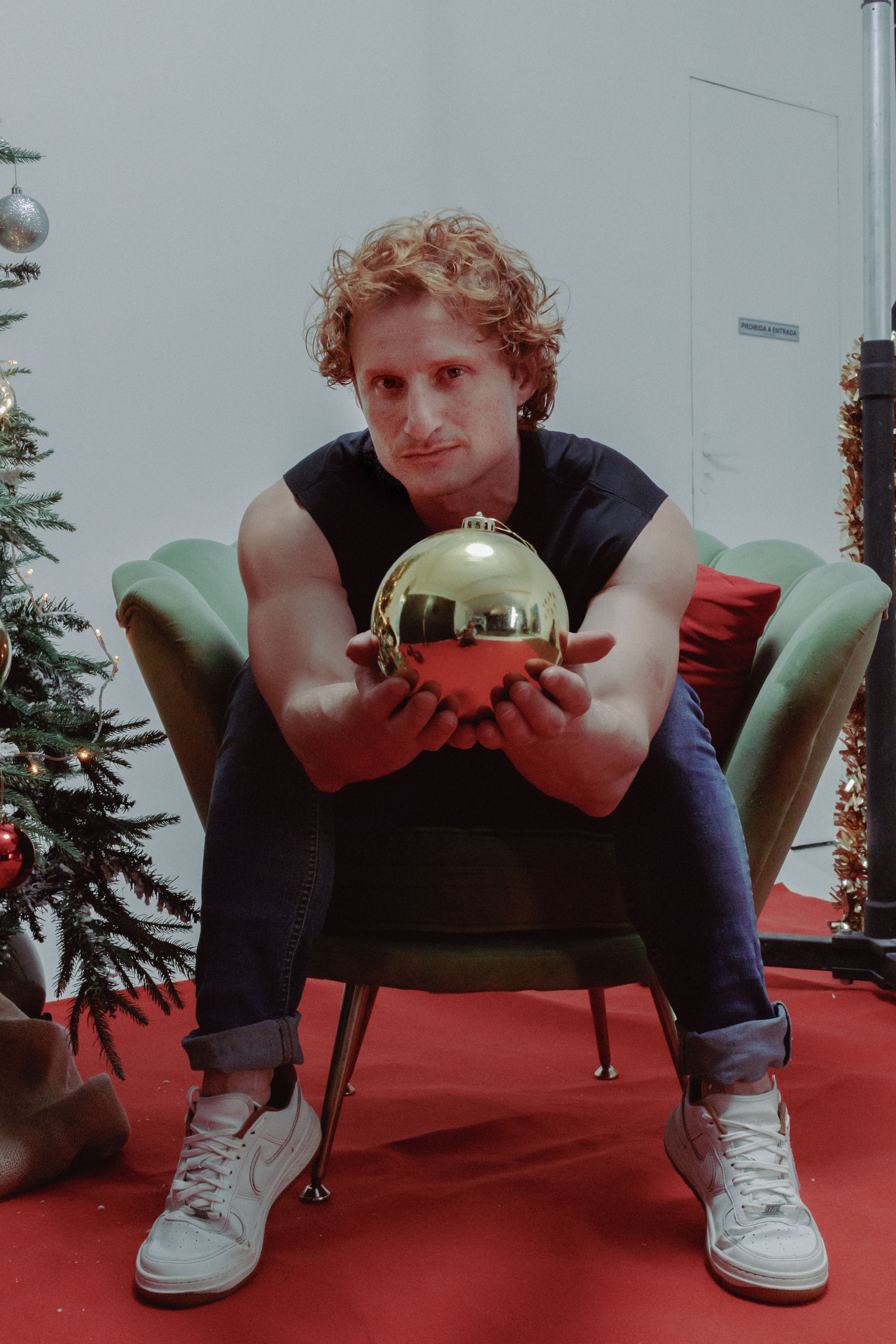 Man sitting on chair holding a golden Christmas ornament, festive setting indoors.