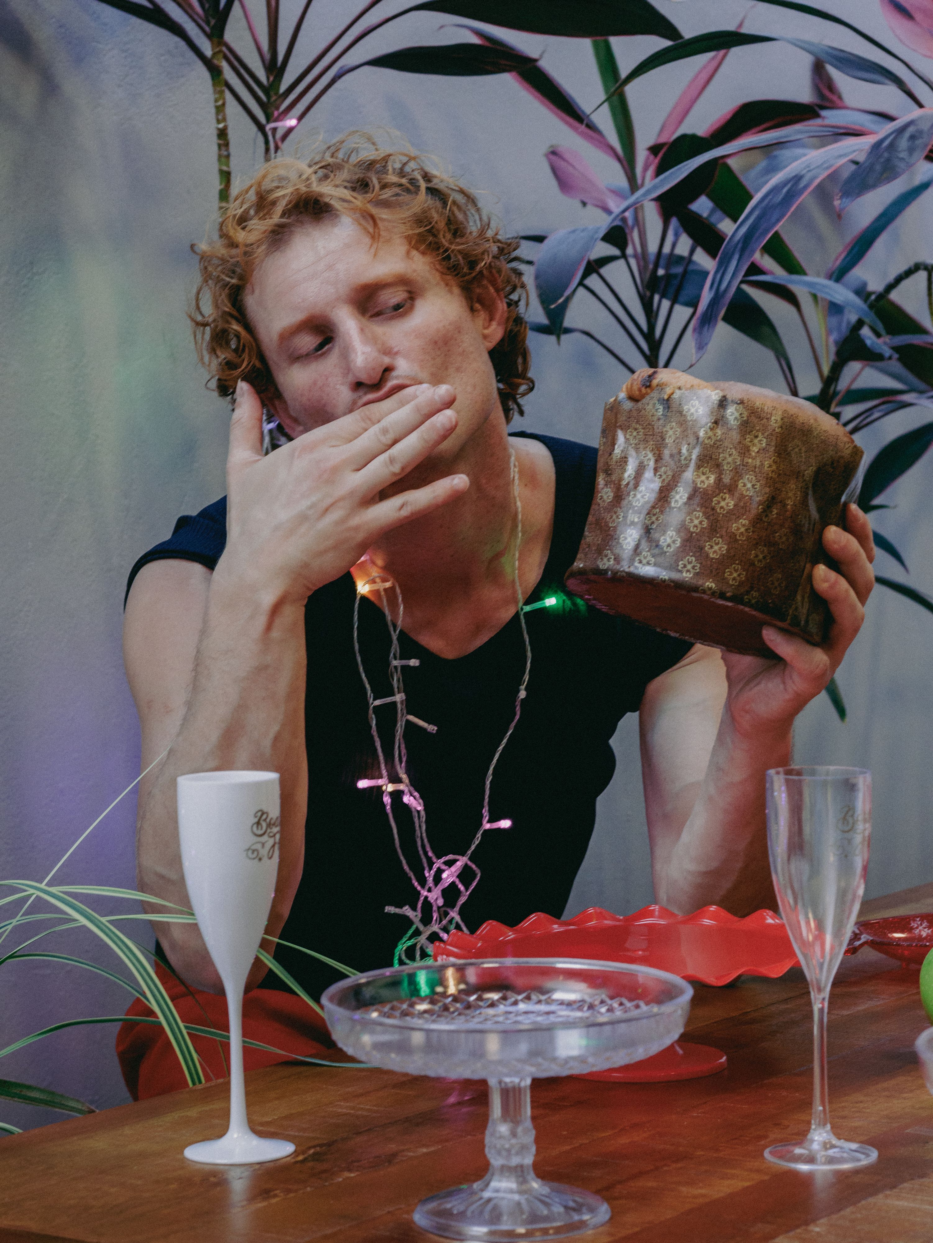 Man sitting at decorated table enjoying cake, surrounded by indoor plants and empty glasses.