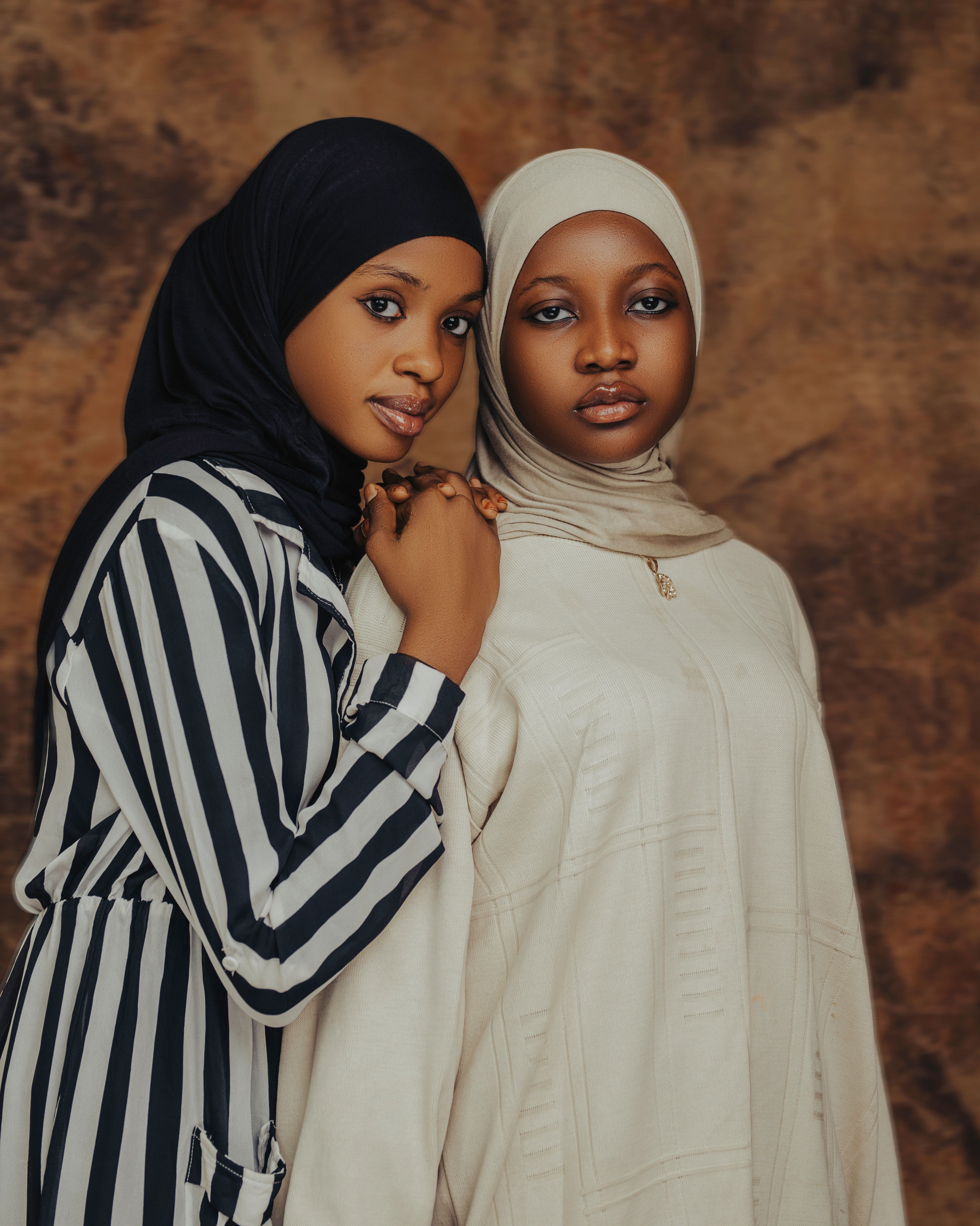 Two young women wearing hijabs pose confidently against a brown backdrop.