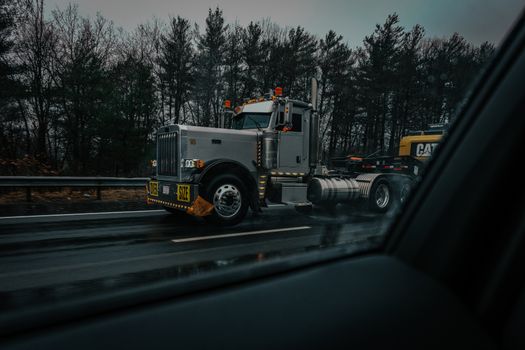 A large truck with an oversize load on a rainy highway, captured through a car window with dark, moody tones.