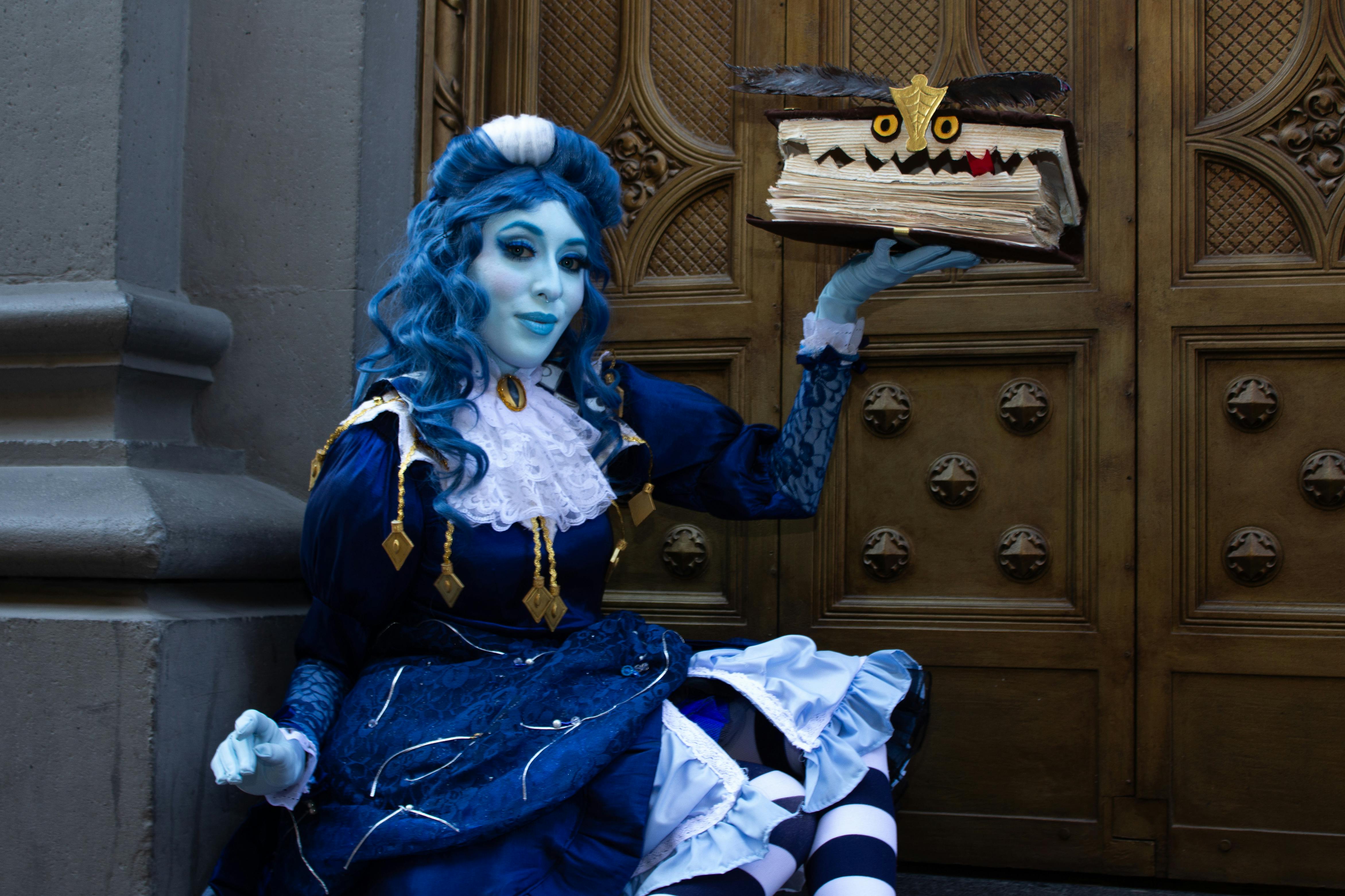 Fantasy cosplayer in blue dress poses with magical book against ornate wooden door.