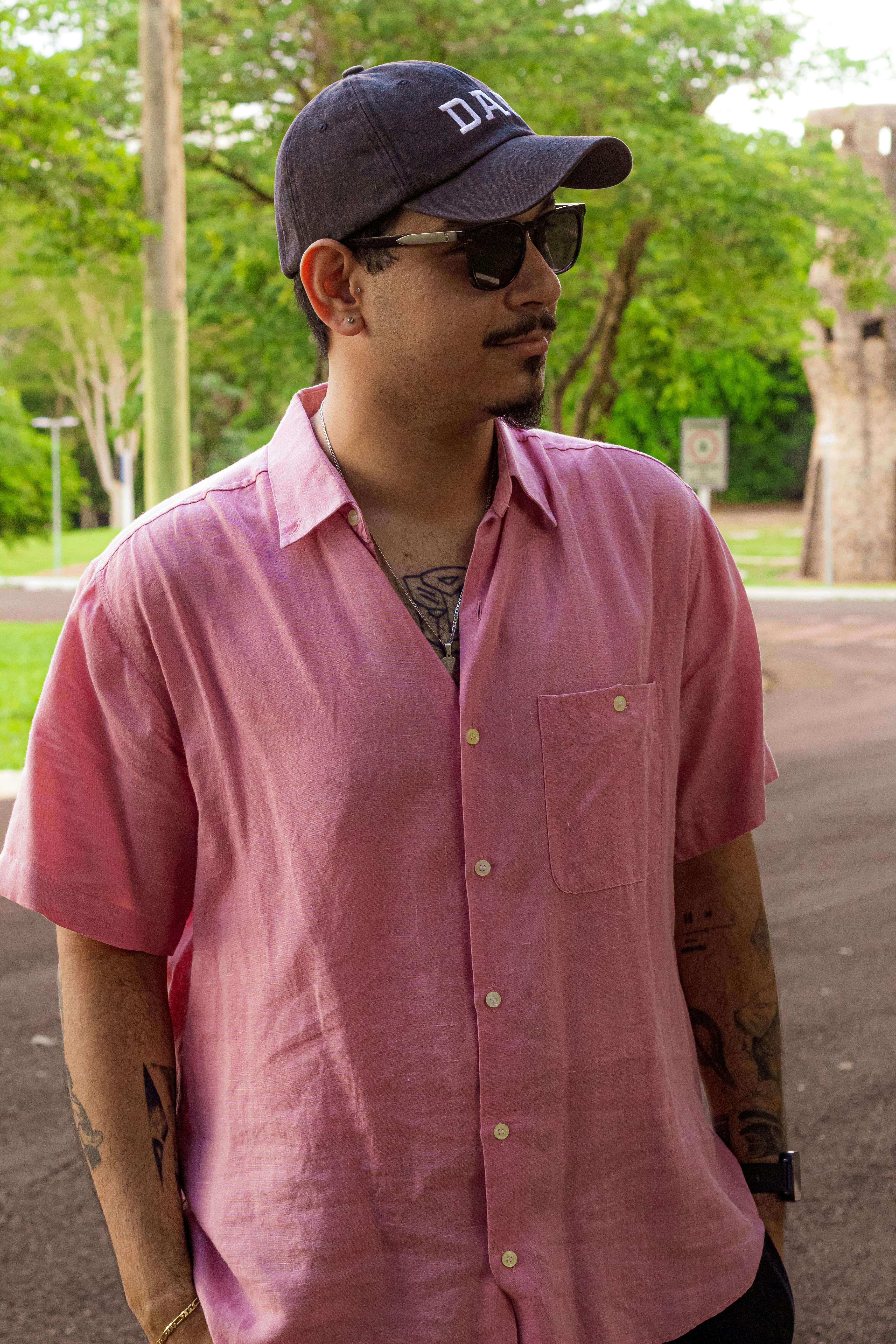 Man in pink shirt and cap in a lush green setting, exuding casual style.