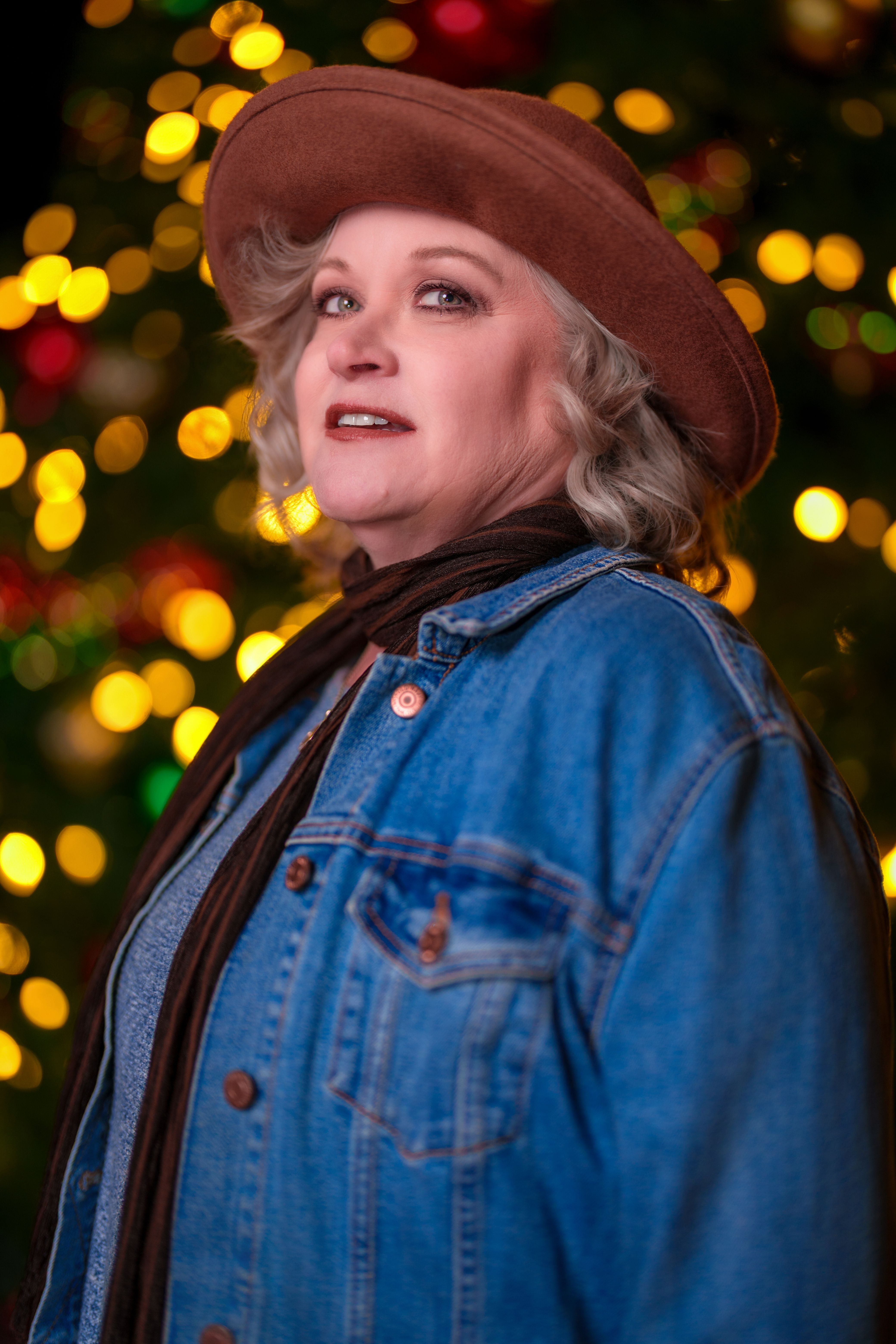 Woman in denim and hat smiles with festive bokeh lights in the background.
