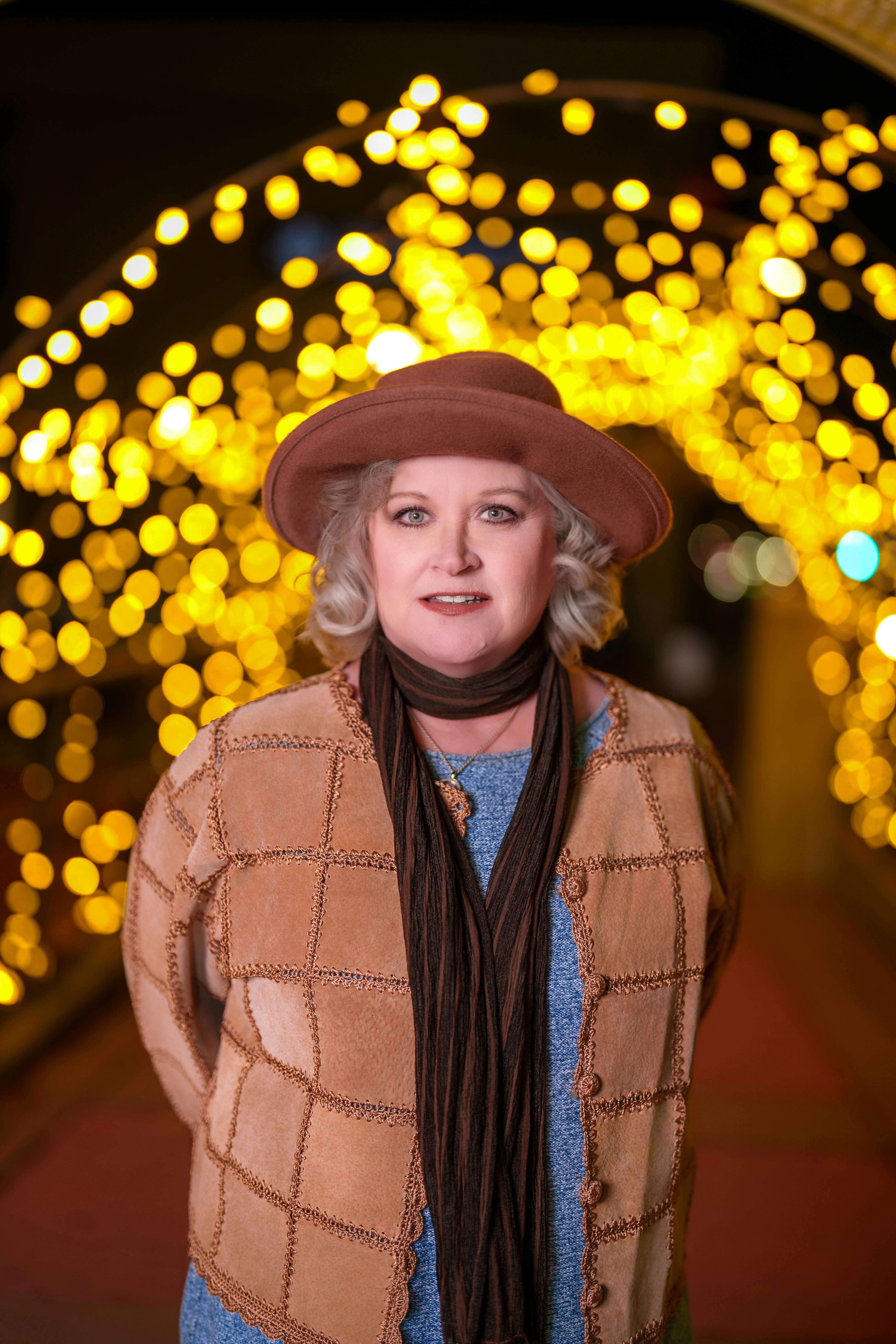 A fashionable woman in a hat poses in front of glowing bokeh lights at night.
