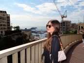 Smiling woman on a bridge overlooking a scenic Mediterranean harbor with cruise ships and urban buildings in the background.