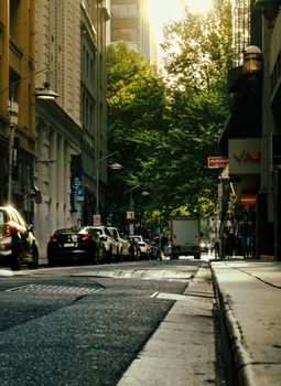 City street view with cars and people, buildings lining the road, and trees in the background.
