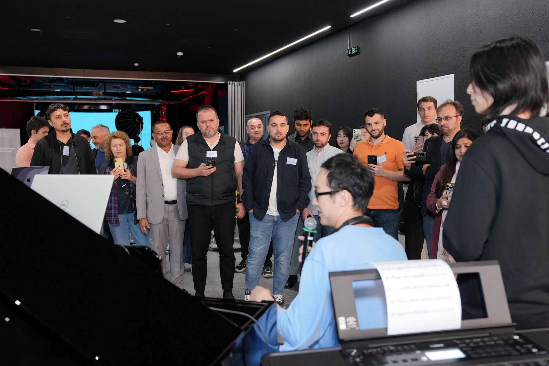 A diverse group of adults attentively watching a piano demonstration indoors. Related image for 2024 Europe Smart Hotels Innovation Day.