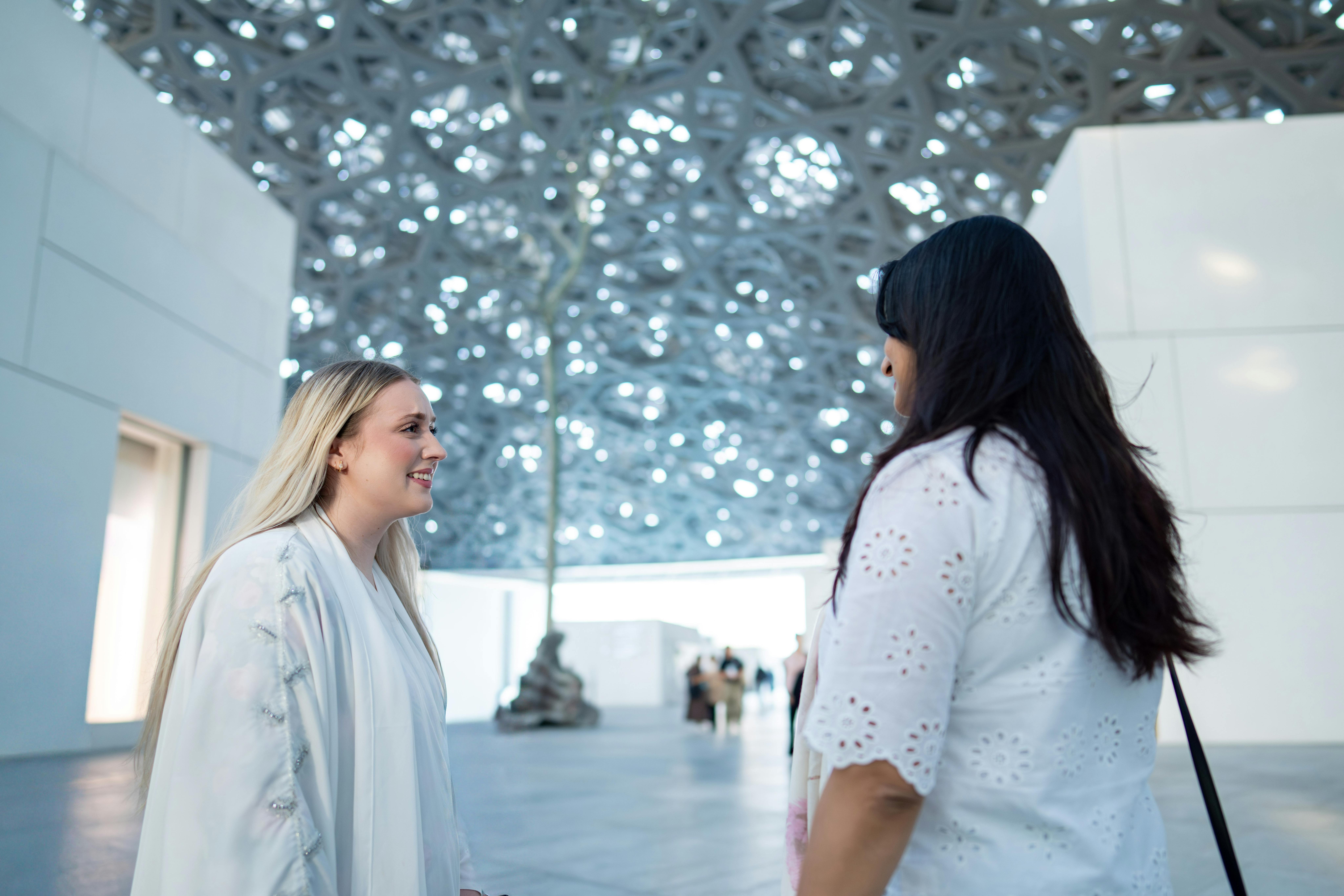 Two women enjoying conversation under the iconic Louvre Abu Dhabi architectural dome.