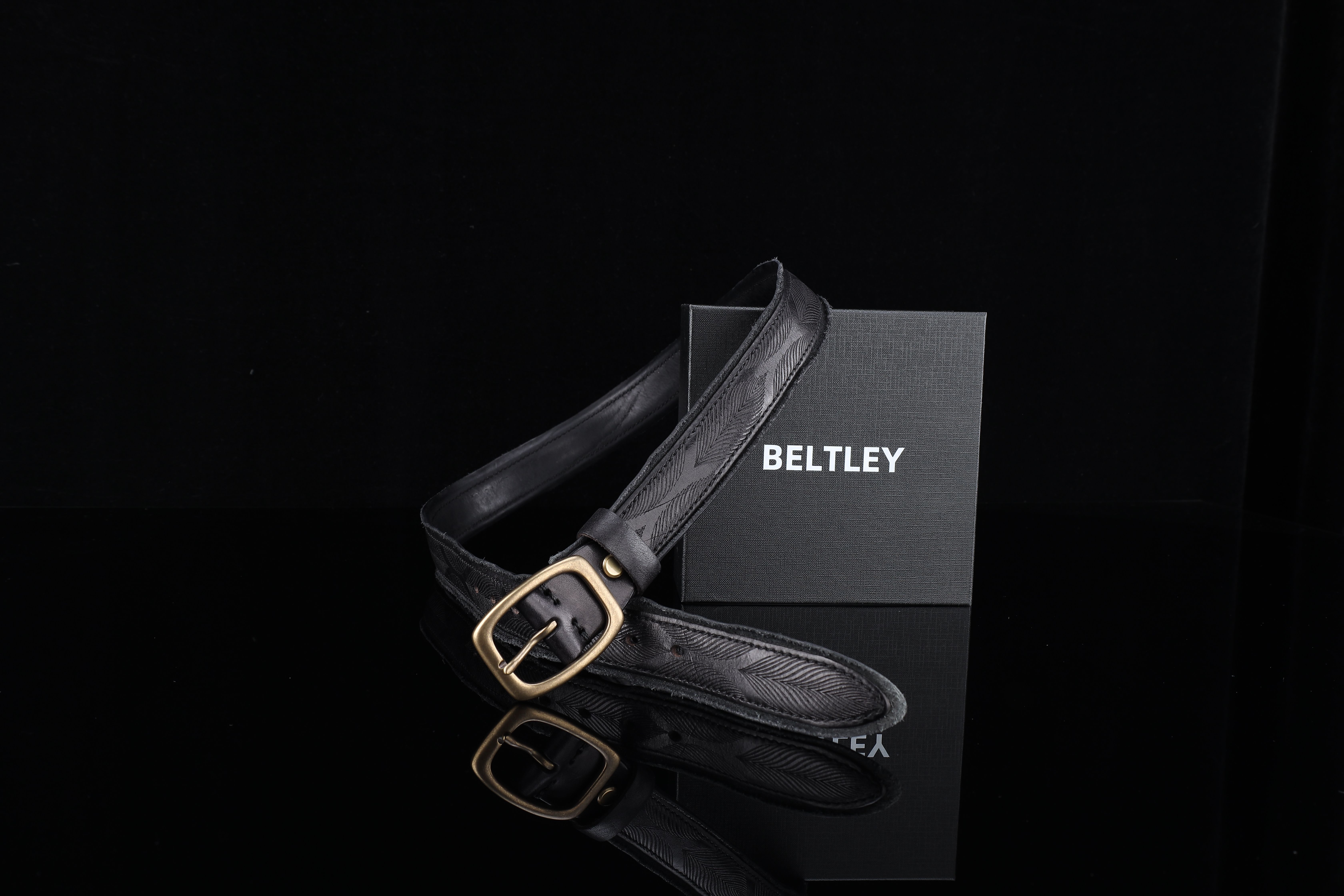 Stylish black leather belt with a golden buckle and box on a sleek reflective surface.
