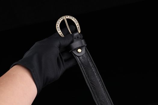 Black leather belt with intricate design and gold buckle, held by a gloved hand against a dark background.