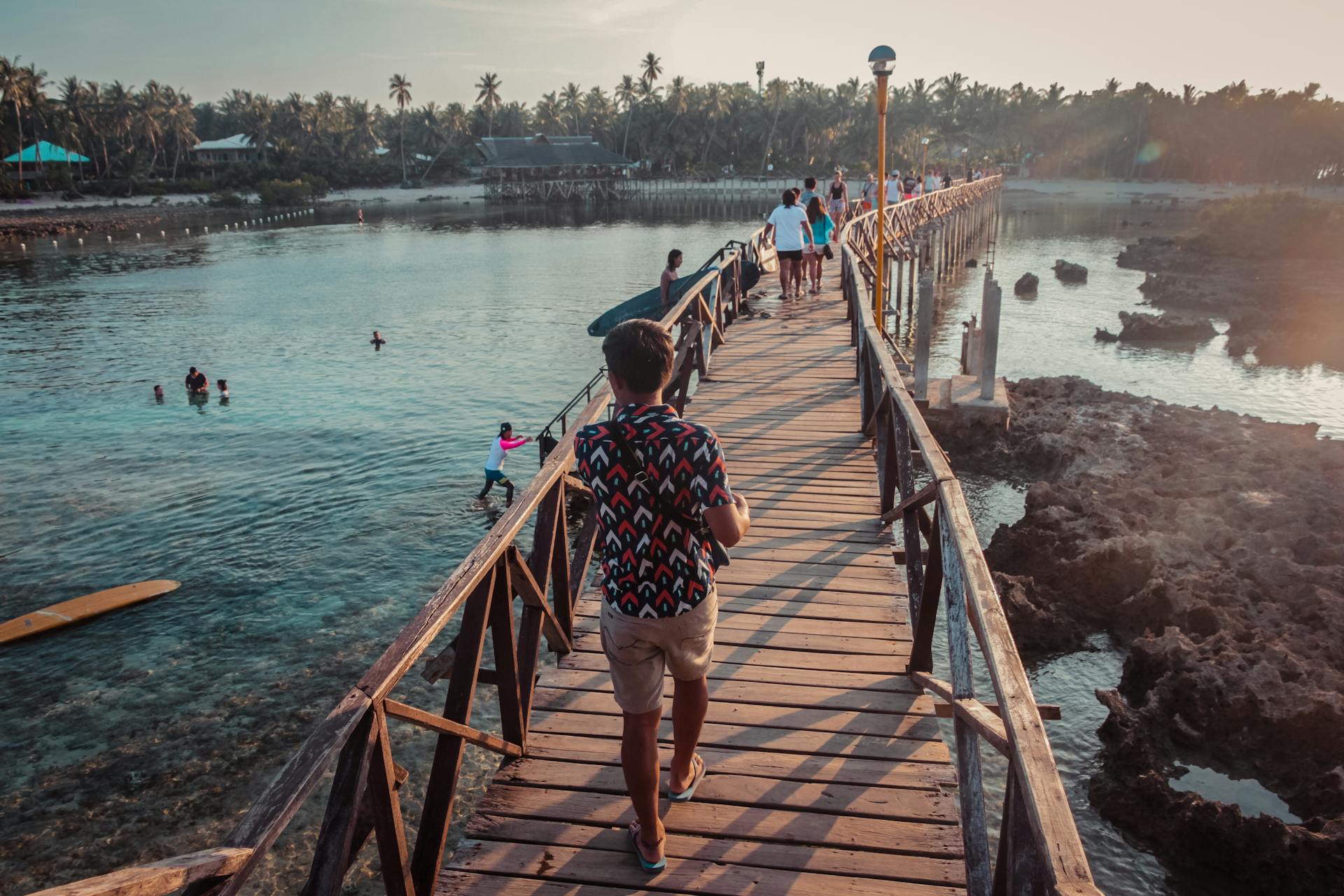 People walk across a scenic wooden bridge in General Luna, Philippines at sunset.