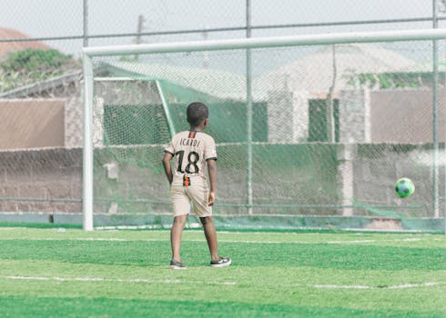 A young boy wearing a jersey stands ready near the soccer goal on a sunny day.