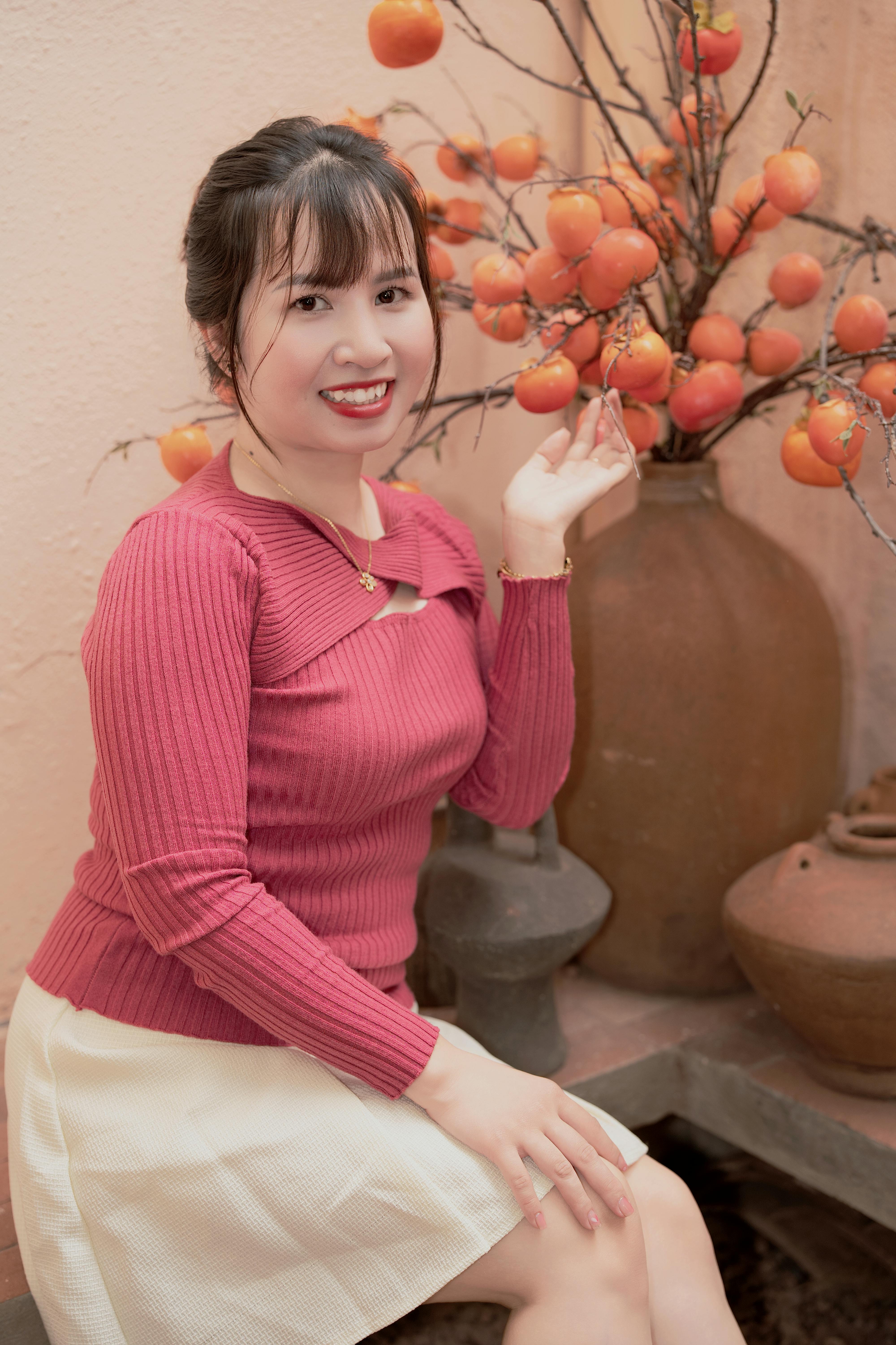 무료 빨간 스웨터를 입은 여성이 실내 감꽃 장식 옆에서 미소 짓고 있어 따뜻한 분위기를 자아낸다. 스톡 사진