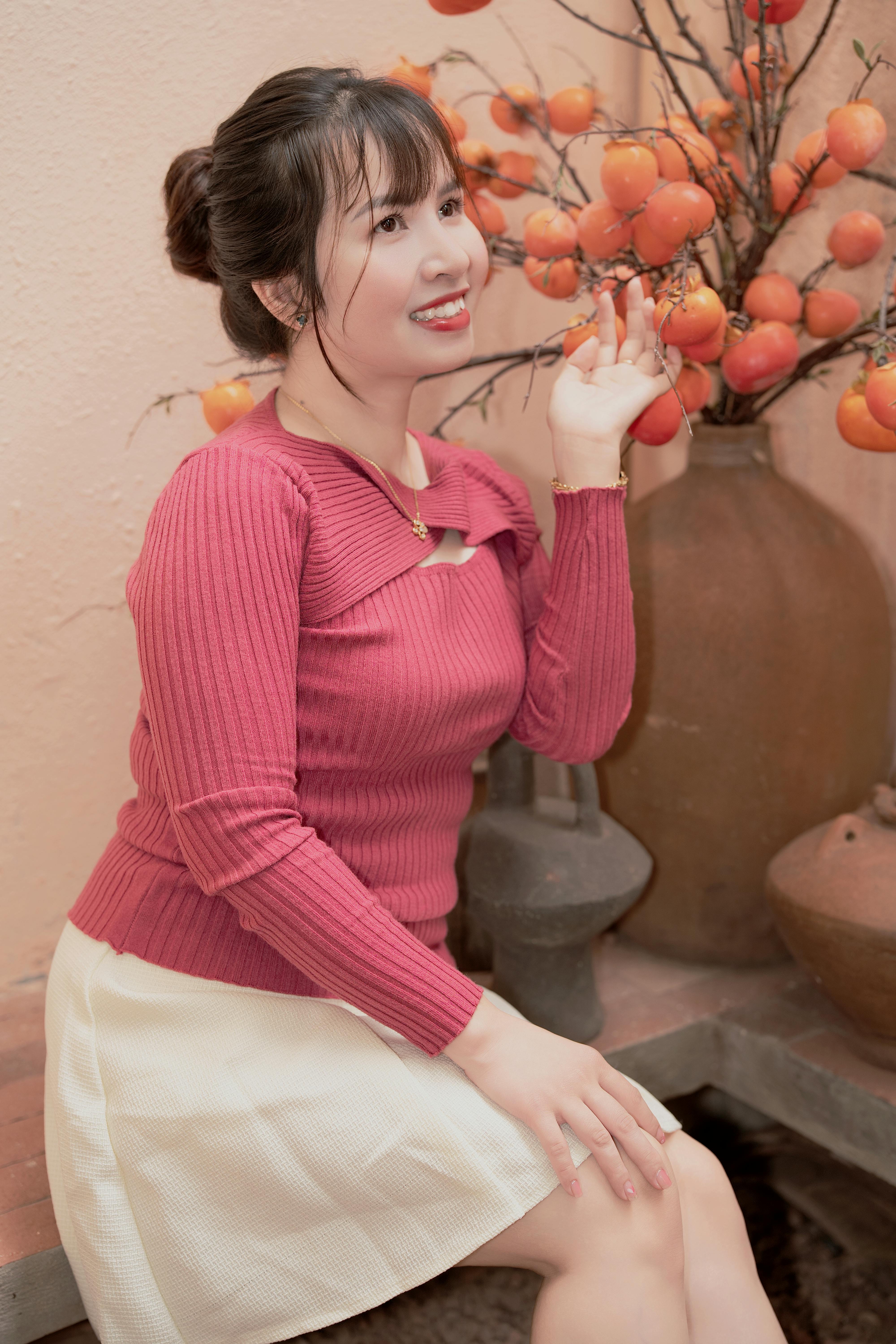 Gratis Seorang wanita bersweater merah memegang buah kesemek di dalam ruangan, menciptakan suasana hangat dan khas musim gugur. Foto Stok