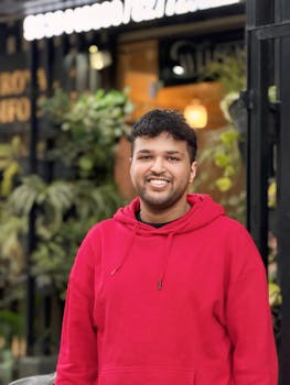 A cheerful person in a red hoodie stands outdoors near a plant-decorated wall.