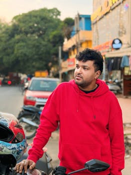 Casually dressed man in red hoodie standing by motorbike on urban street corner.
