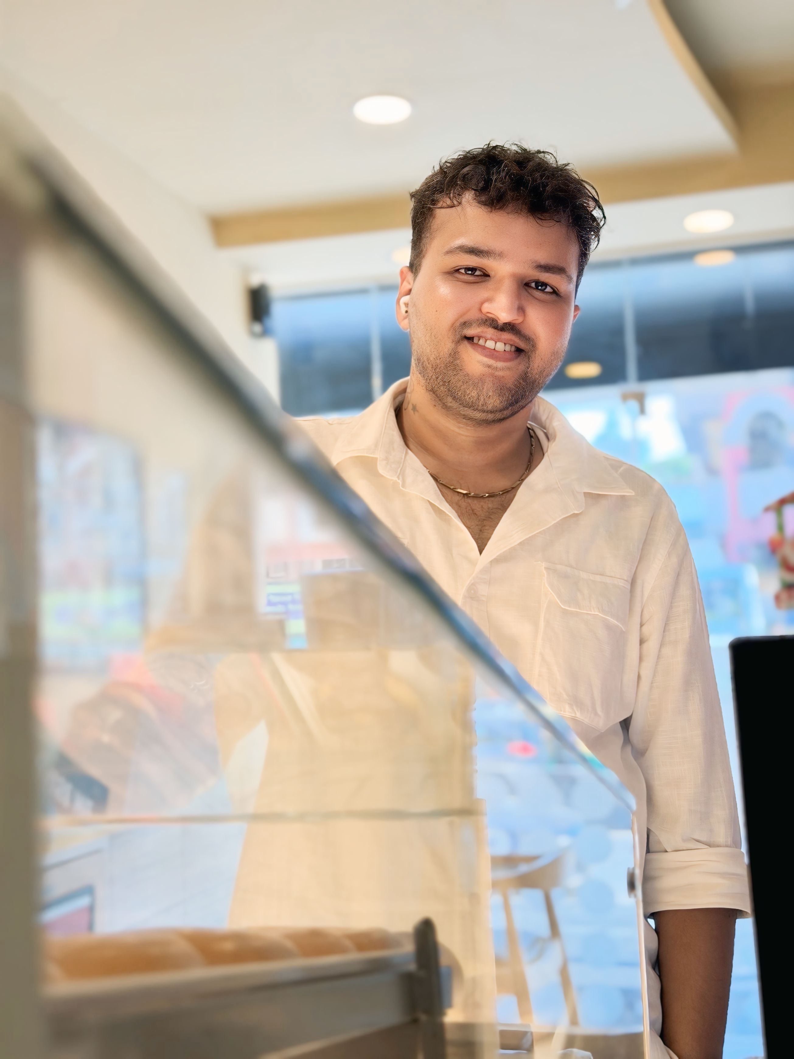 A barista in a white shirt smiles warmly inside a cafe, creating a welcoming atmosphere.