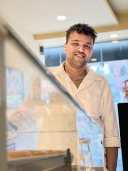 A barista in a white shirt smiles warmly inside a cafe, creating a welcoming atmosphere.