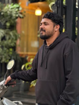 Man in black hoodie standing beside a scooter in a green urban environment.